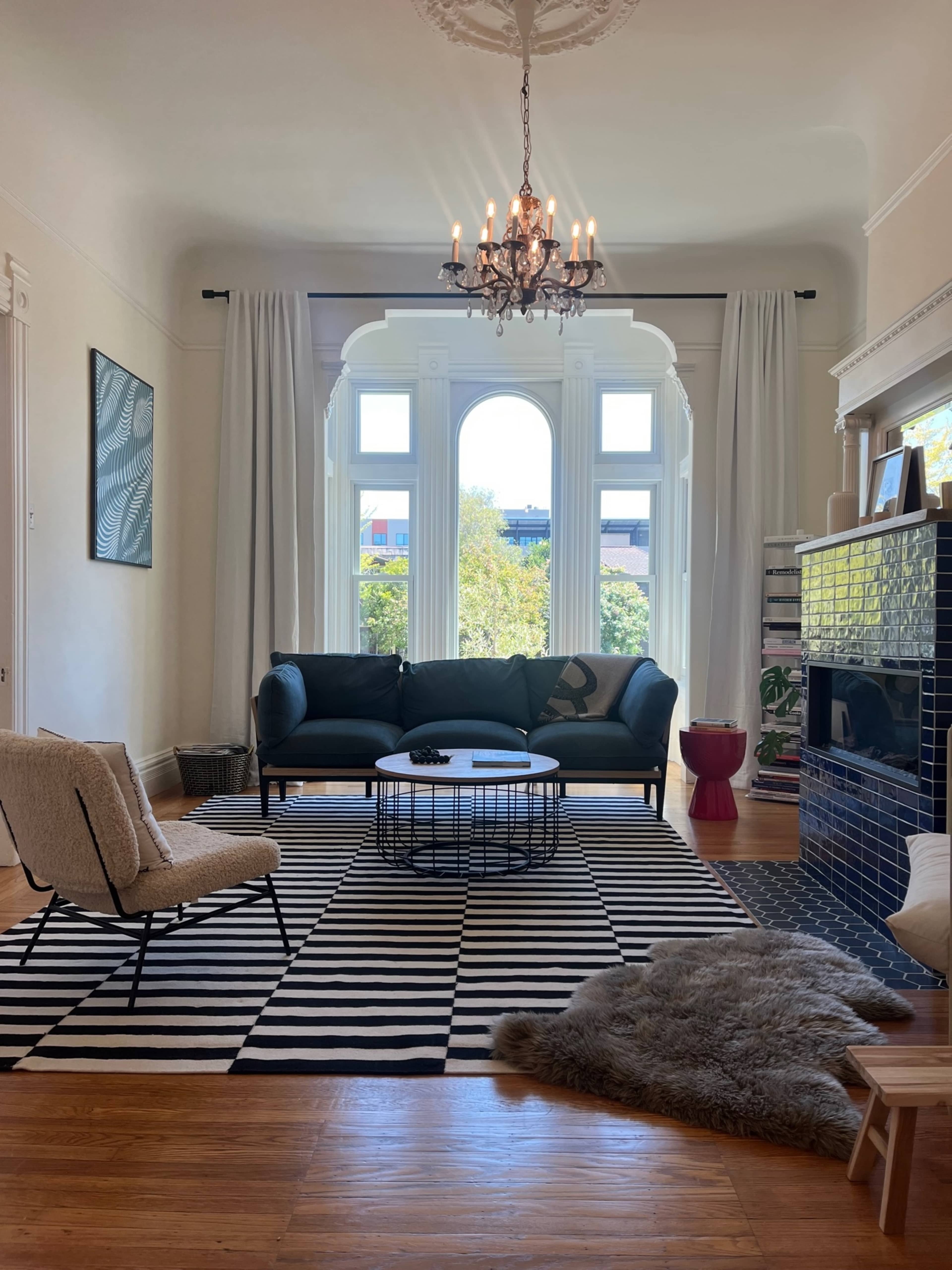 The image shows a well-lit living room featuring a green sofa, a round coffee table on a striped rug, and a decorative fireplace with a blue tile surround.