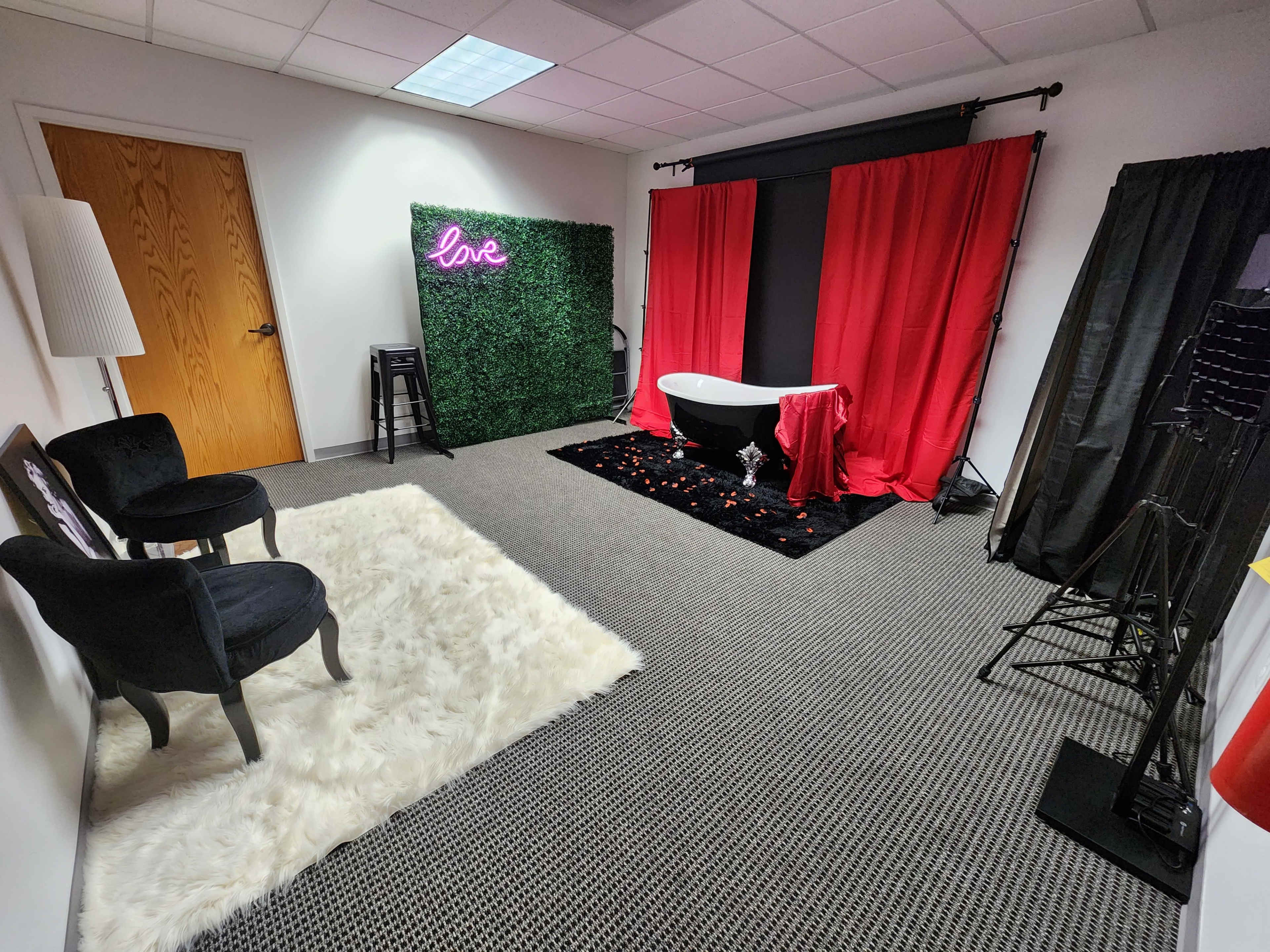 The scene features a styled indoor room with a bathtub draped in red fabric, surrounded by decorative elements like a faux green wall, black chairs, and a fluffy white rug.