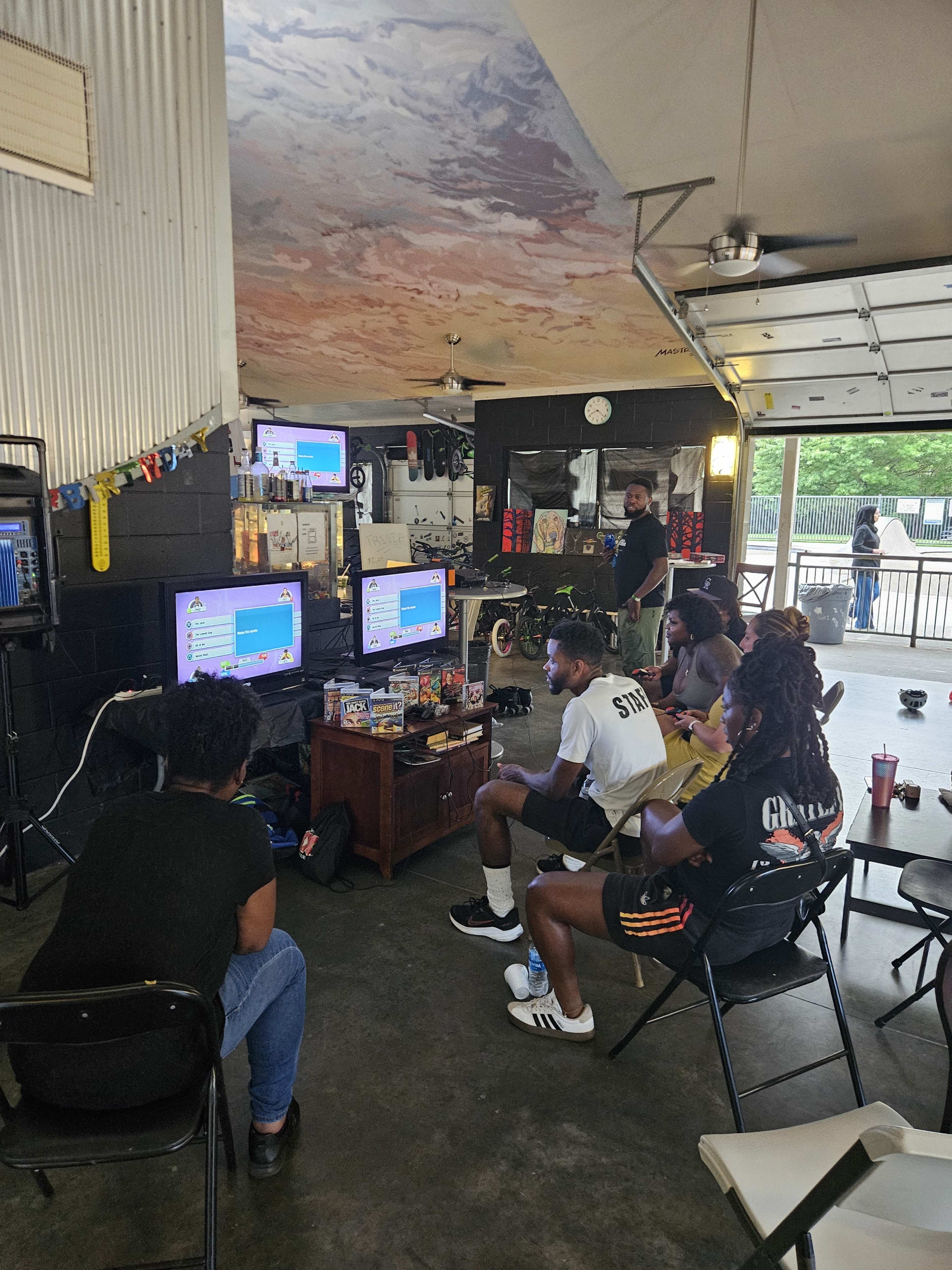 A group of five people are seated in a garage, watching multiple screens displaying video games.