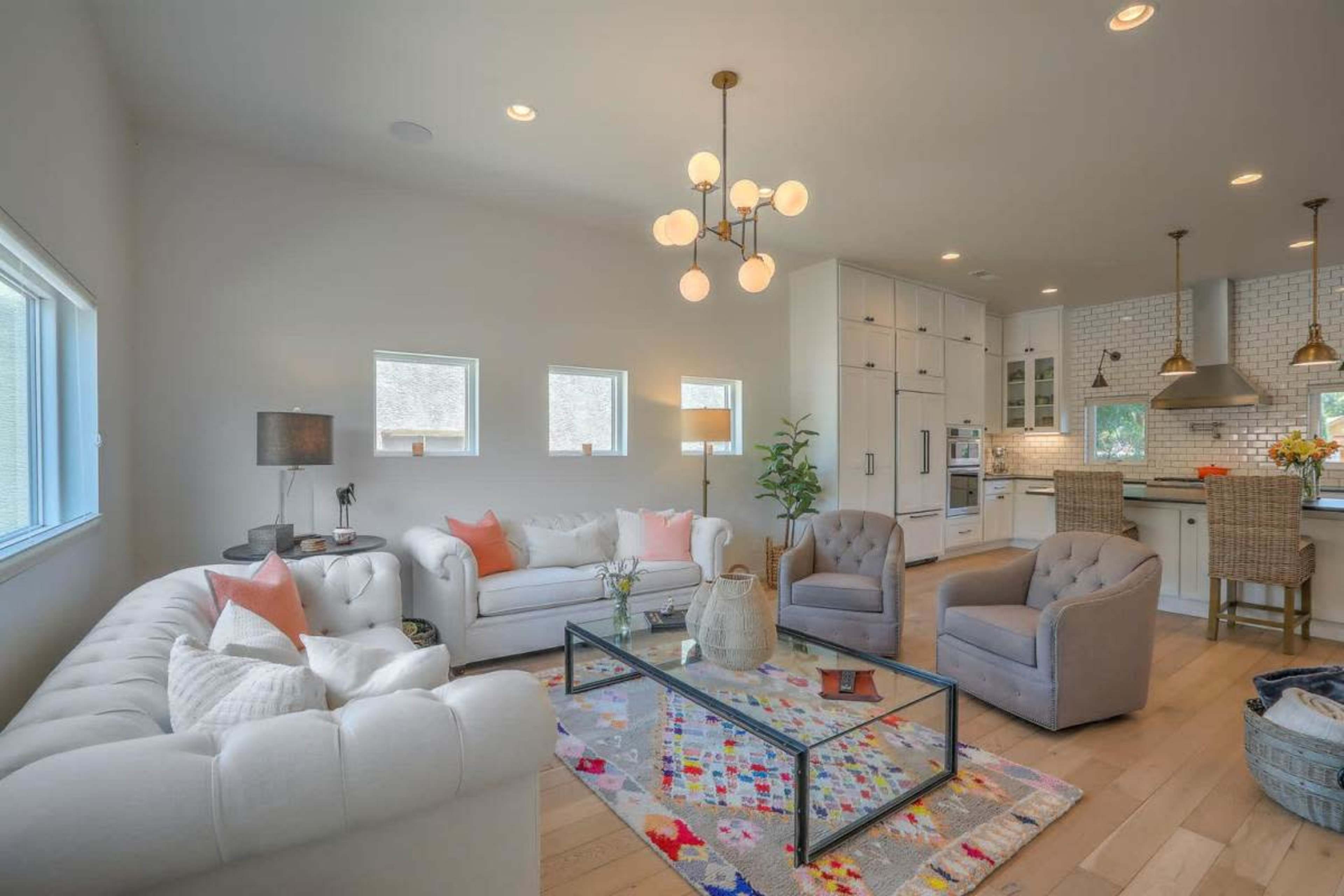 The image shows a modern living room featuring a white sectional sofa, light gray armchairs, a glass coffee table, and a colorful area rug, illuminated by a statement chandelier.