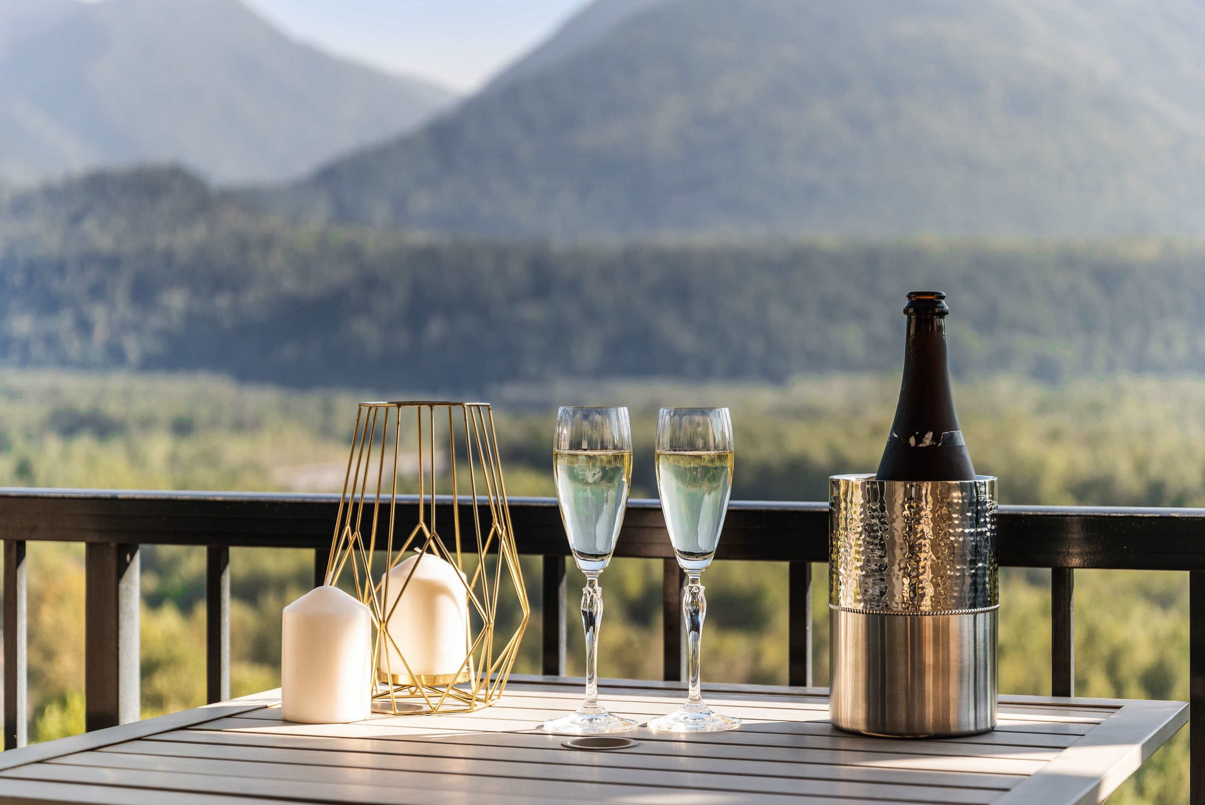 A bottle of sparkling wine sits in an ice bucket beside two filled champagne glasses on a wooden table with a scenic mountain backdrop.