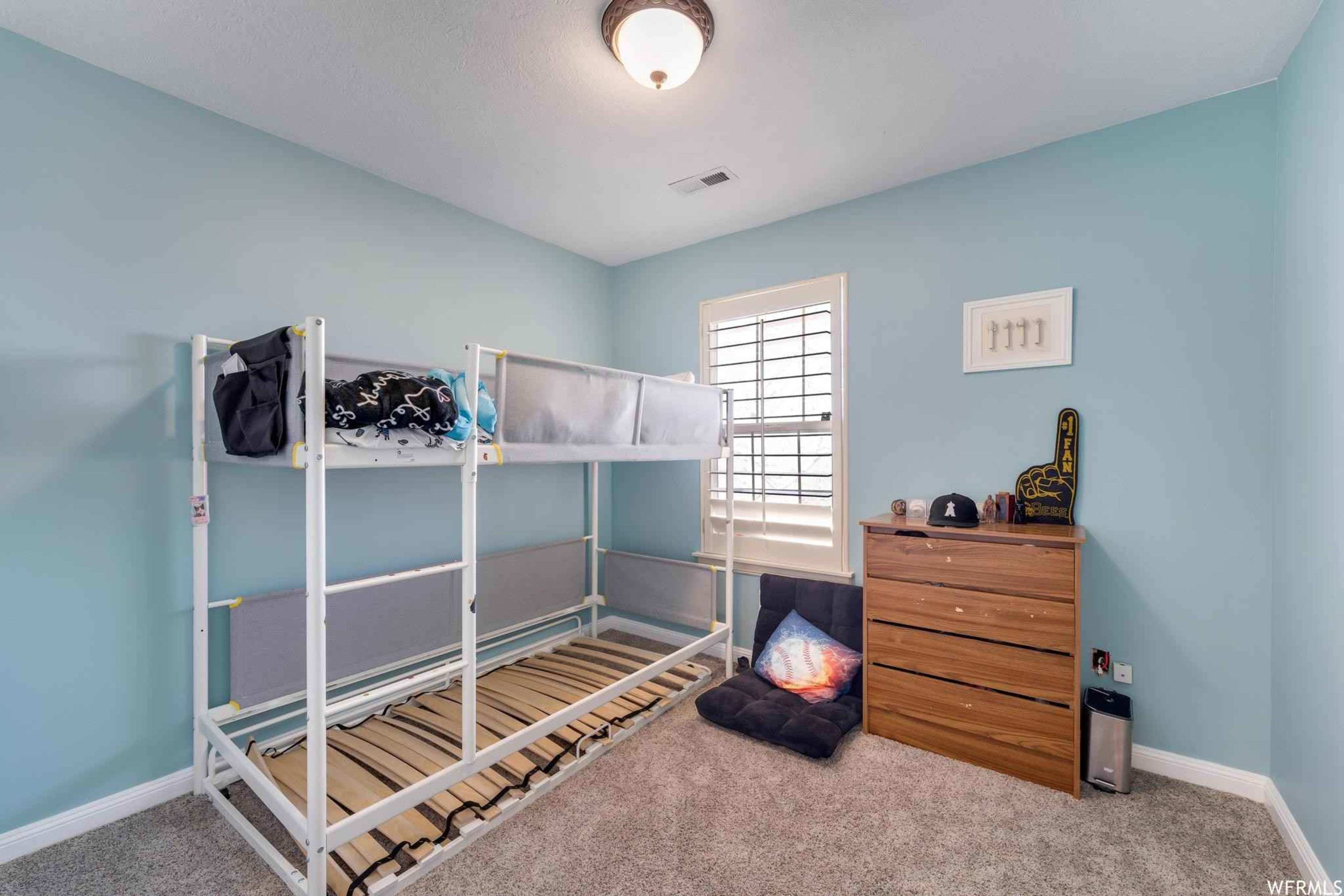 A light blue bedroom with a lofted bed frame, a wooden dresser, and a cozy seating area with a black cushion on a carpeted floor.