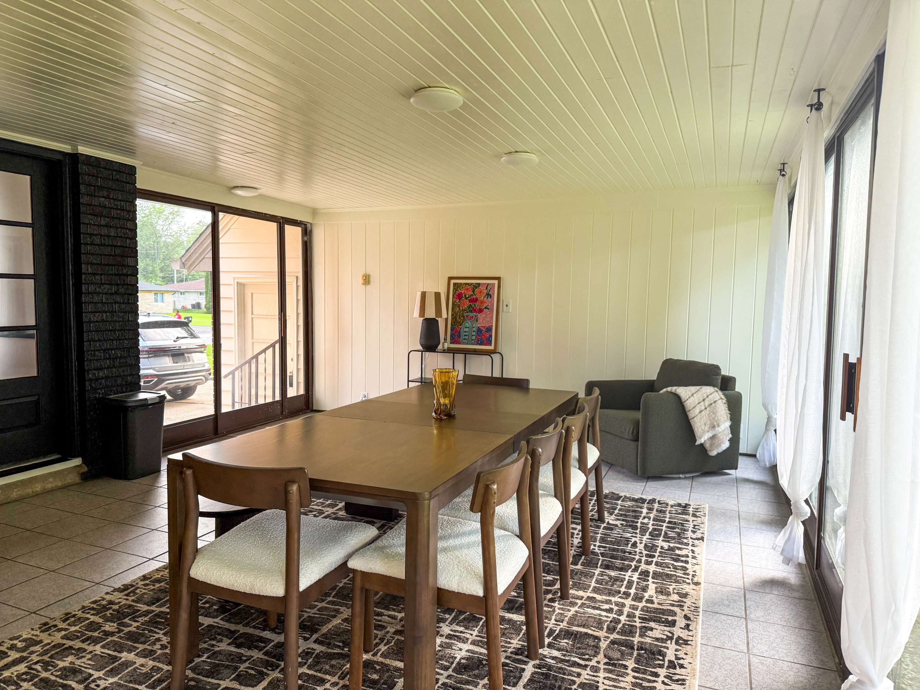 The image shows a spacious, brightly lit dining area with a wooden table and chairs, a patterned rug, and a cozy seating area in the corner.