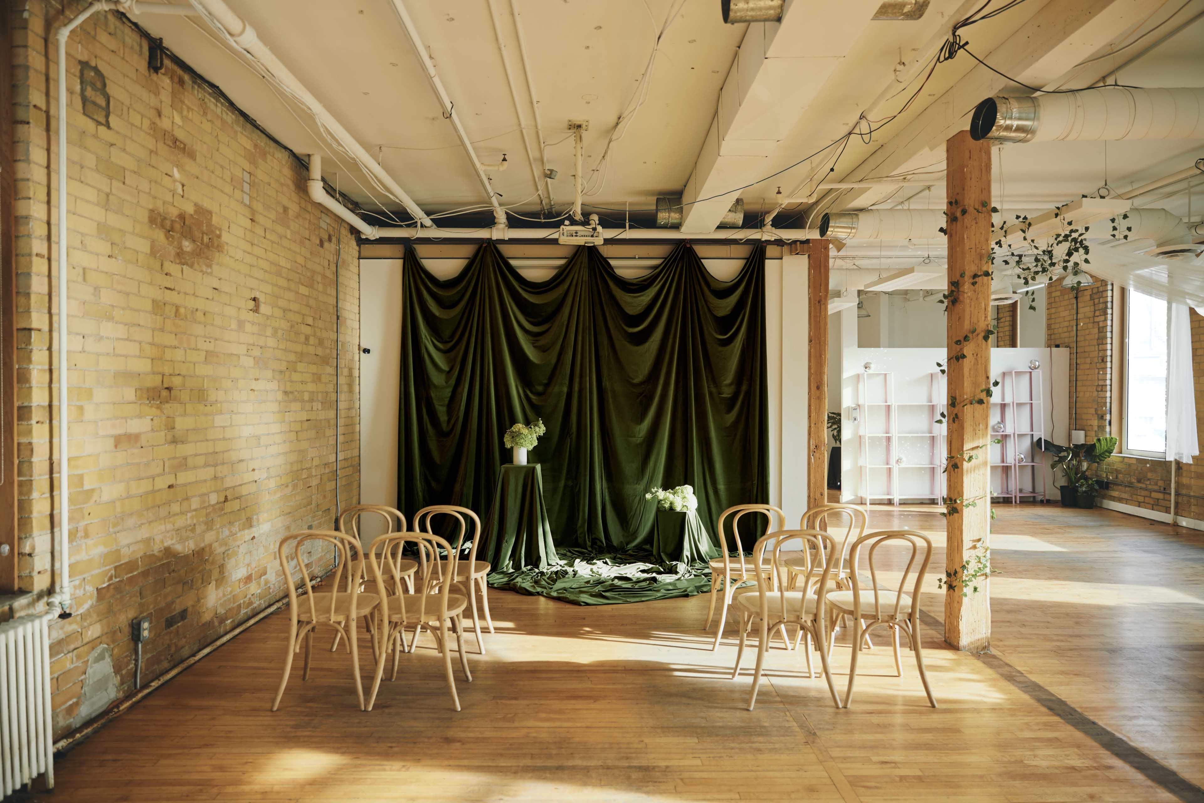 The image shows a spacious room with exposed brick walls and a green velvet backdrop, accompanied by a round table and several wooden chairs arranged in front of it.
