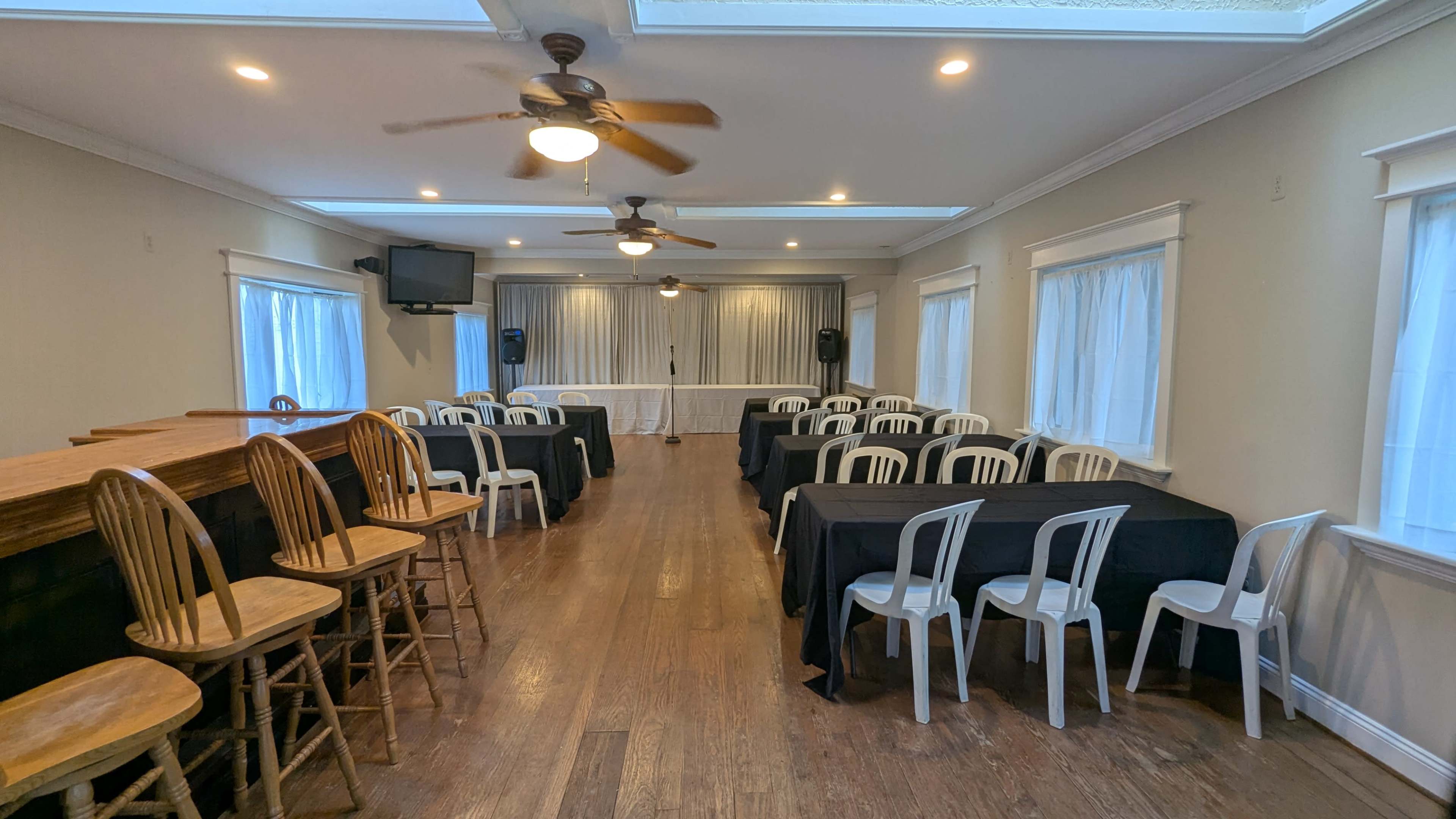 A spacious indoor venue features rows of black-covered tables and white chairs, with a bar setup along one side and a stage area at the front.