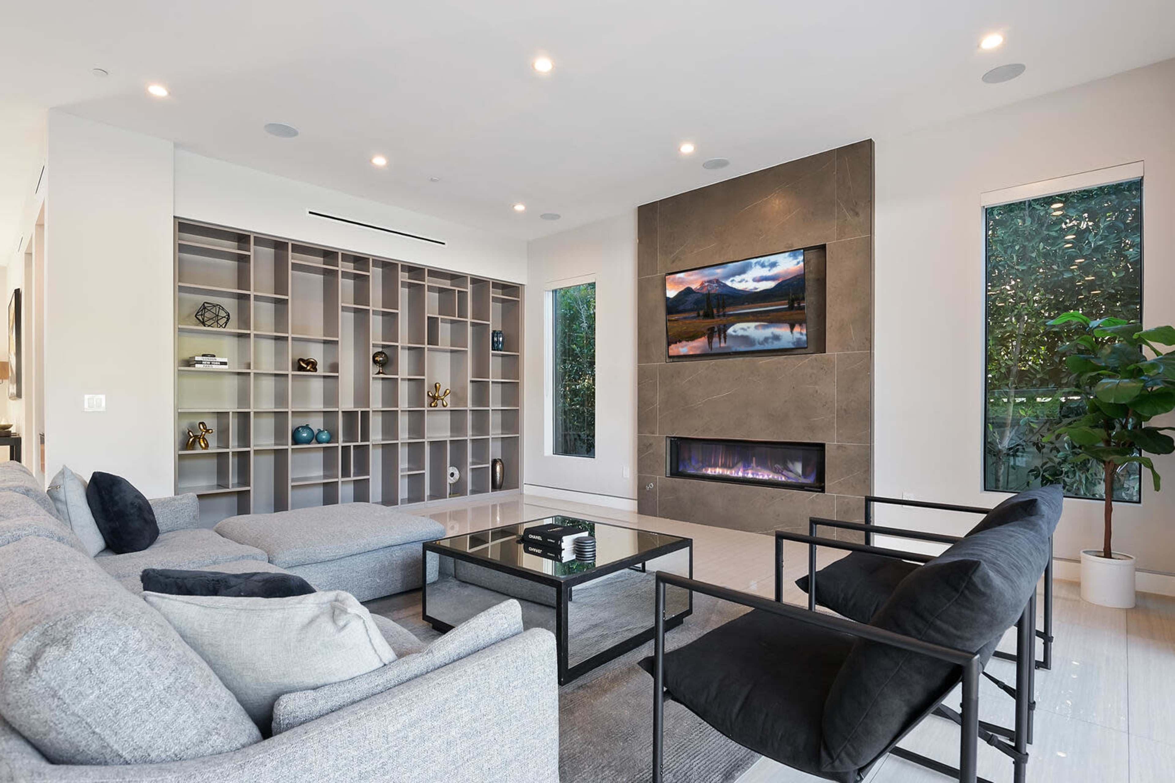 A modern living room features a sectional sofa, two black armchairs, a sleek coffee table, a television mounted above a fireplace, and built-in shelving.