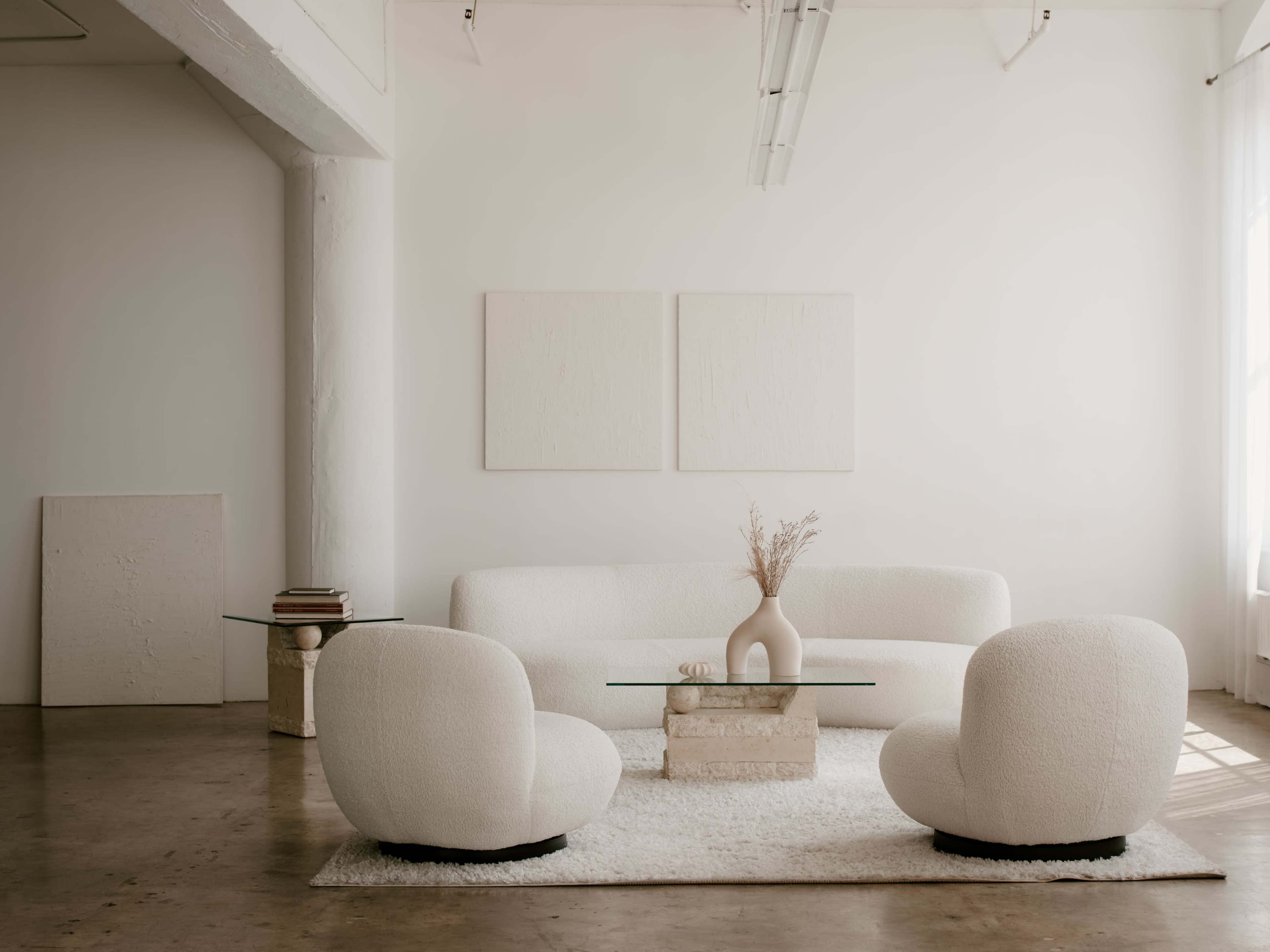 The image shows a minimalist living room with a light-colored sofa, two rounded chairs, a glass coffee table, and a textured white rug.