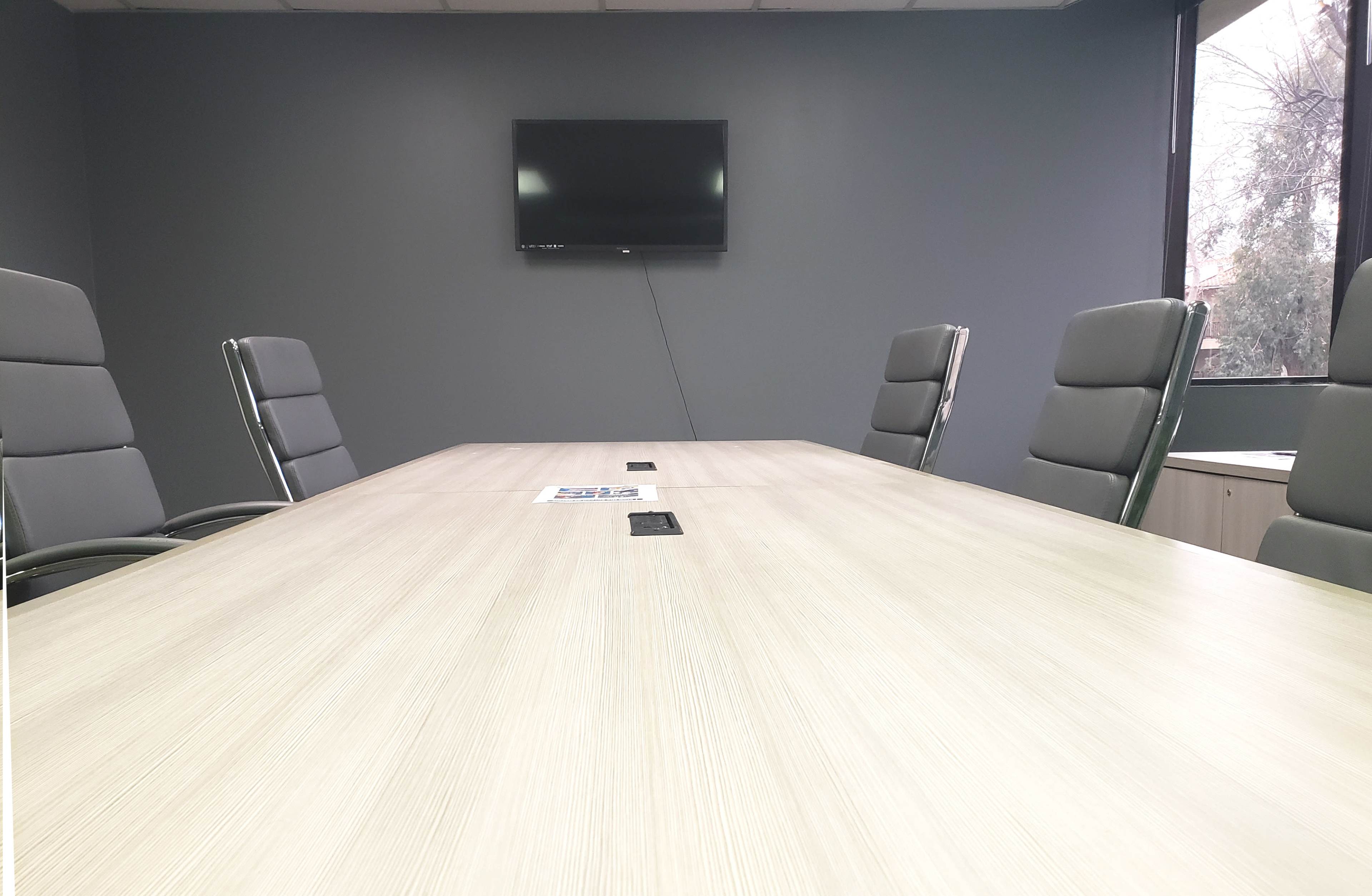 A conference room with a long wooden table, six gray leather chairs, a television mounted on the wall, and large windows letting in natural light.