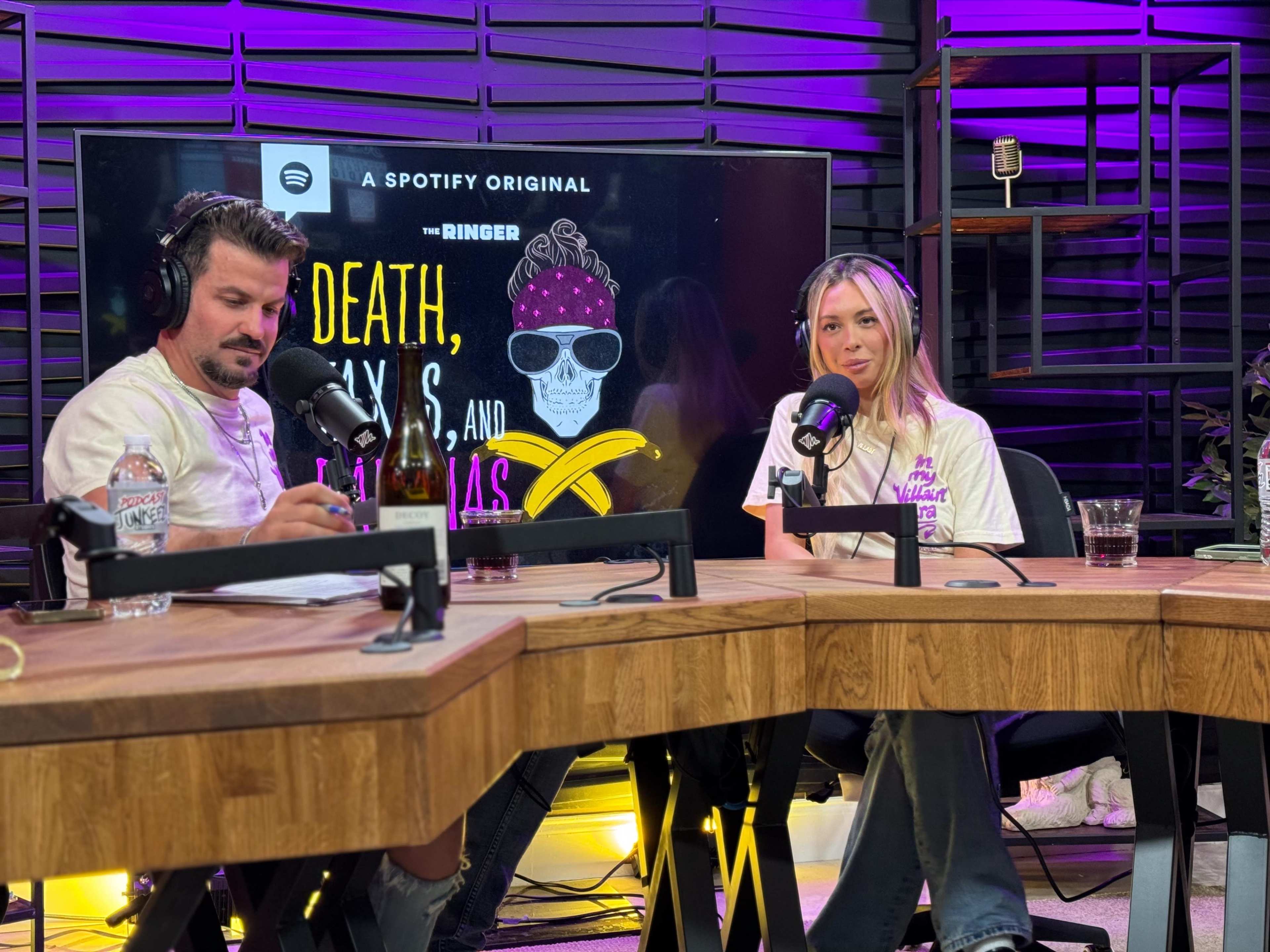 Two individuals sit at a wooden table in a podcast studio, surrounded by microphone setups, a water bottle, and a display featuring a colorful graphic for a Spotify original show.
