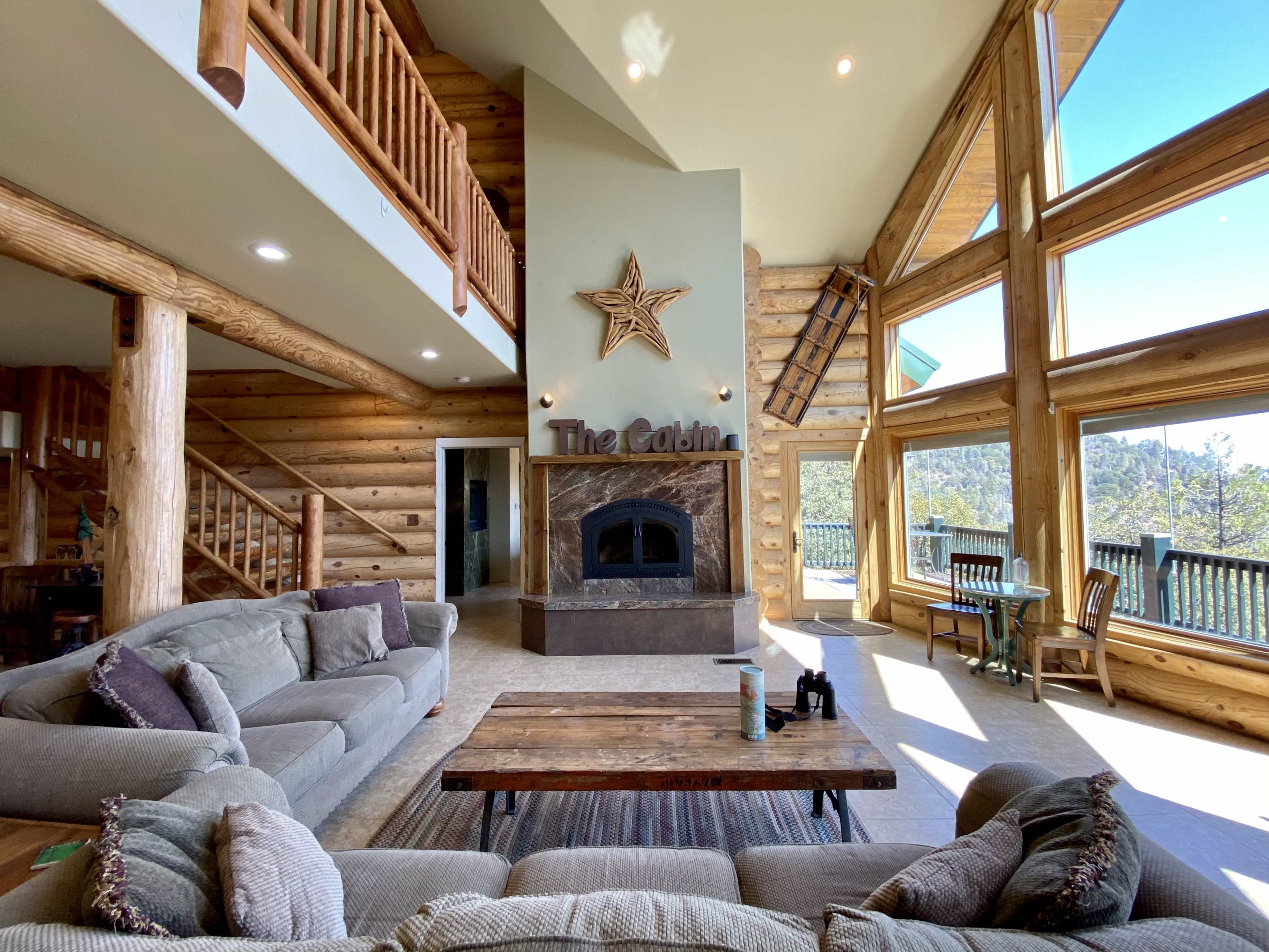 A spacious cabin interior with large windows, a stone fireplace, and wooden beams.