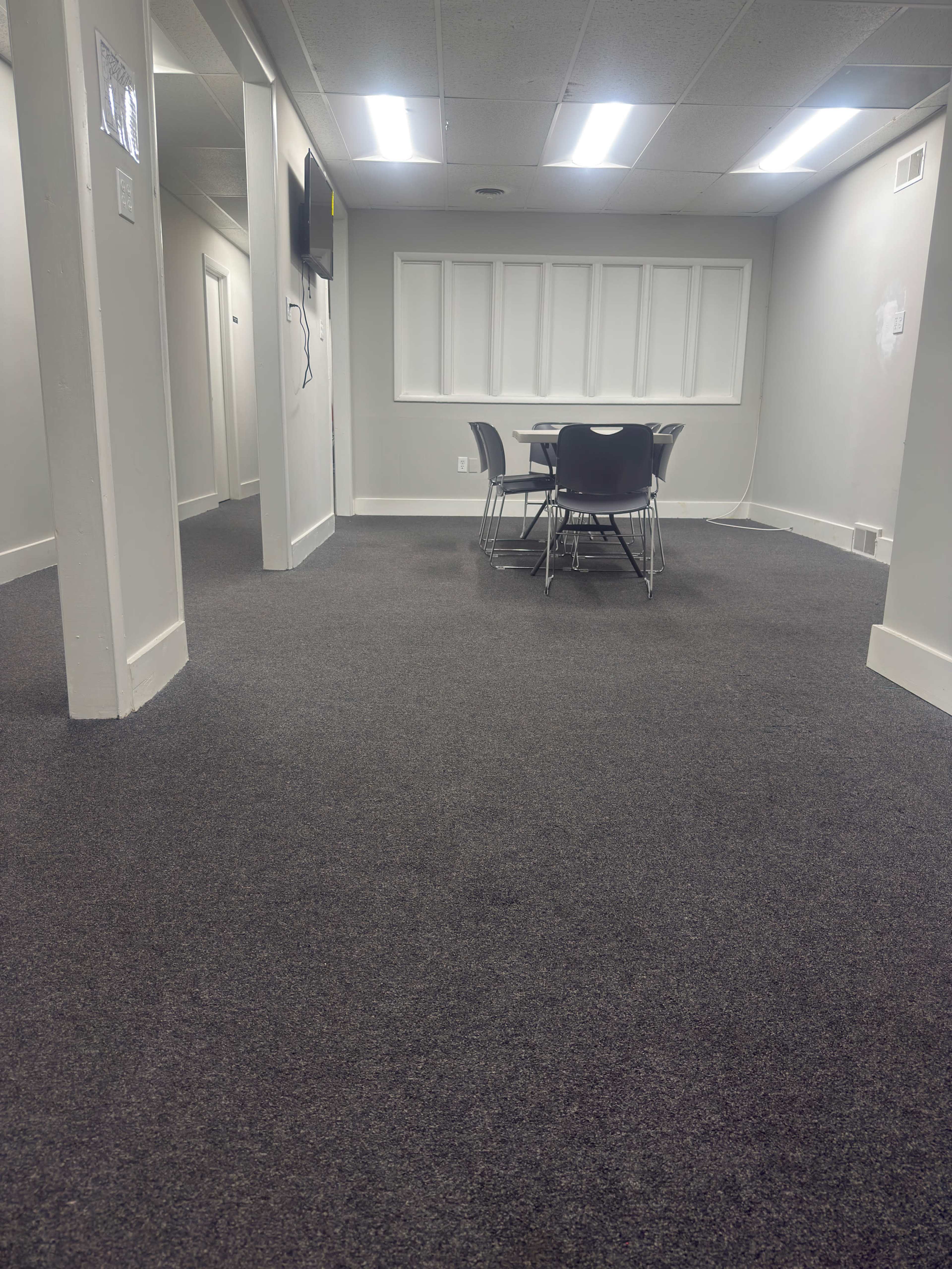 The image shows a carpeted room with a table and chairs in the center, flanked by white walls and lit by overhead fluorescent lights.