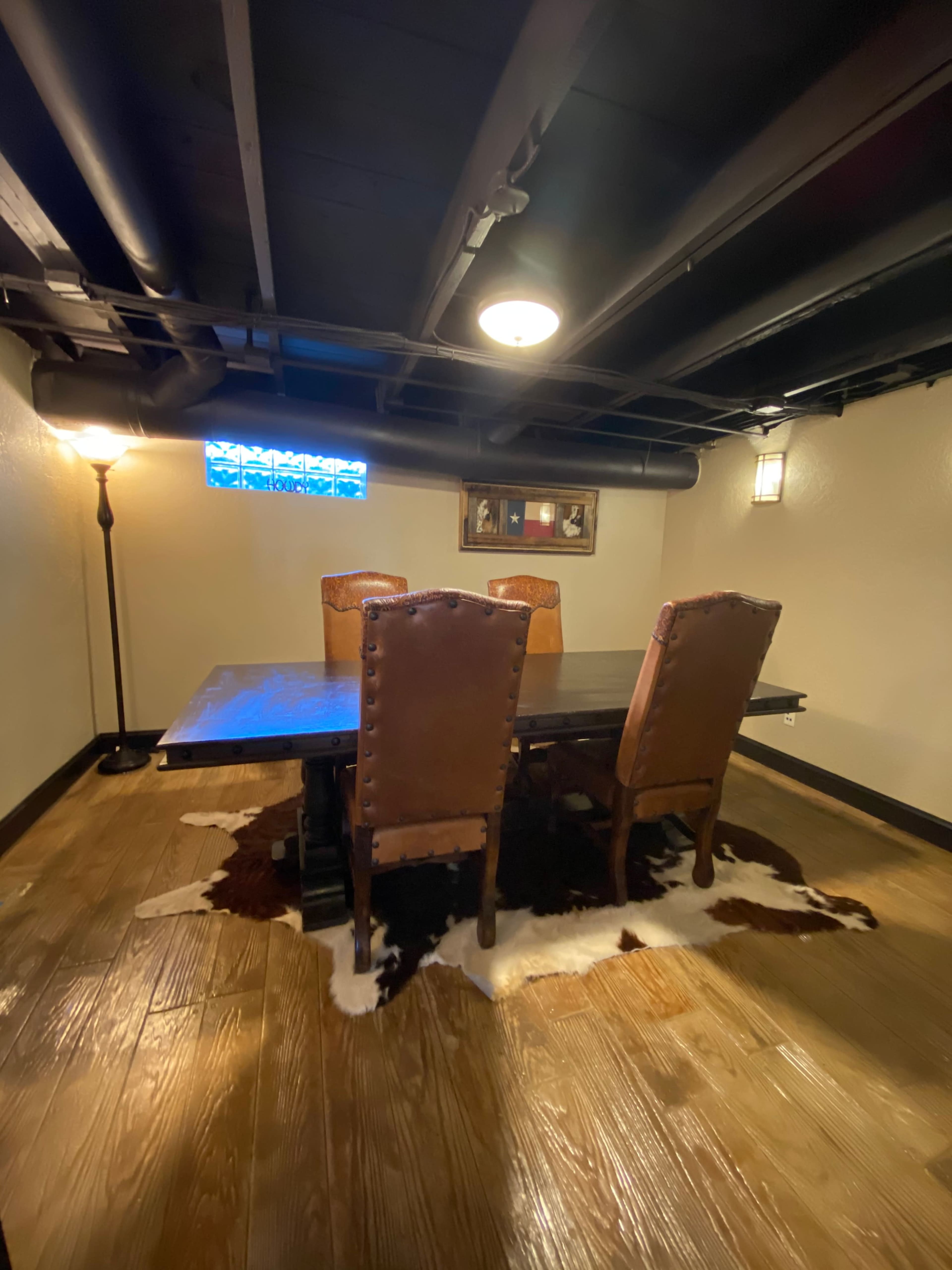 A dimly lit basement dining room with a large wooden table and four high-backed chairs on a wooden floor, accented by a cowhide rug and a small window.