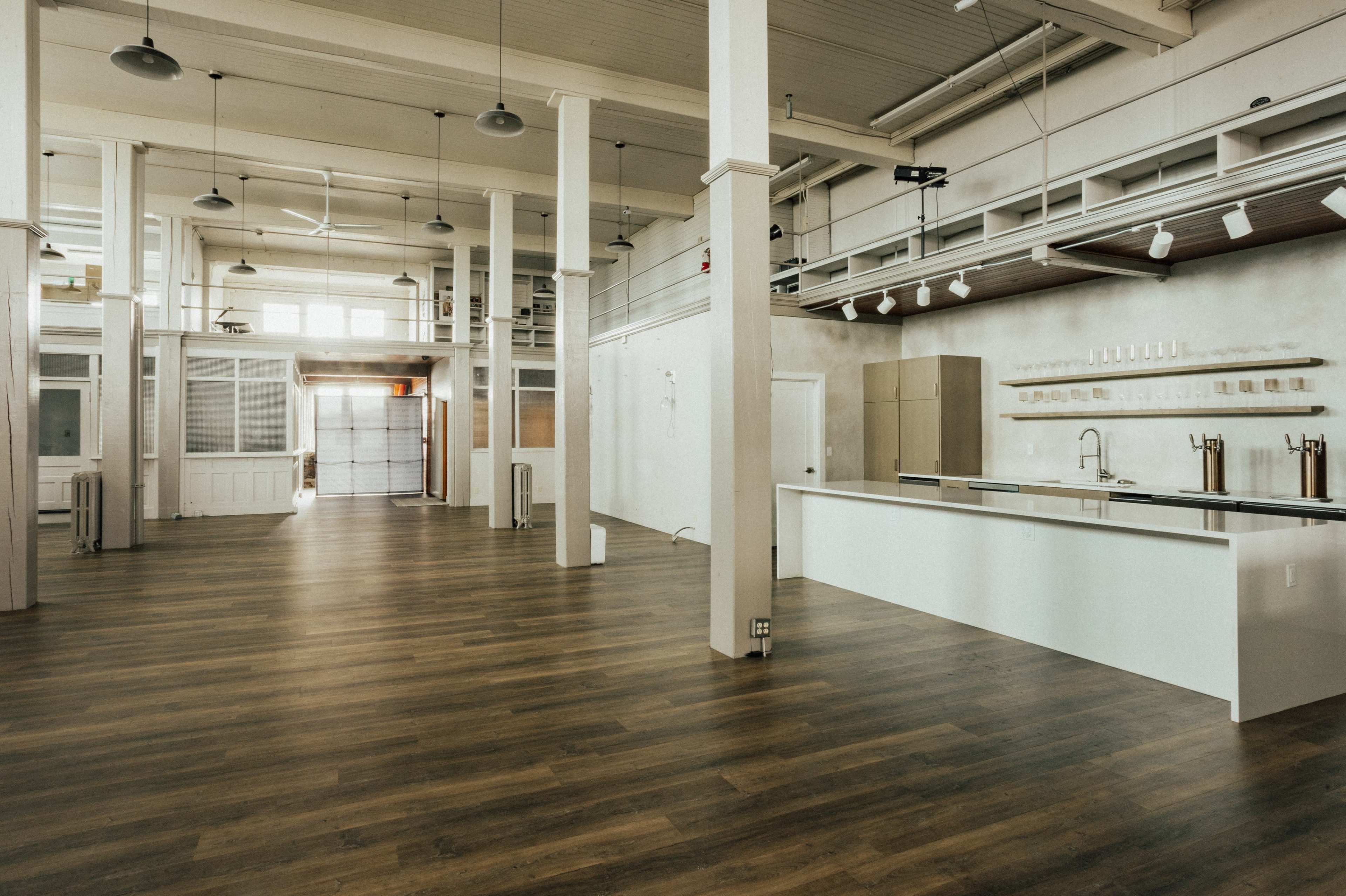 A spacious, modern interior with wooden flooring, high ceilings, and minimalist furnishings, including a long white counter and large windows.