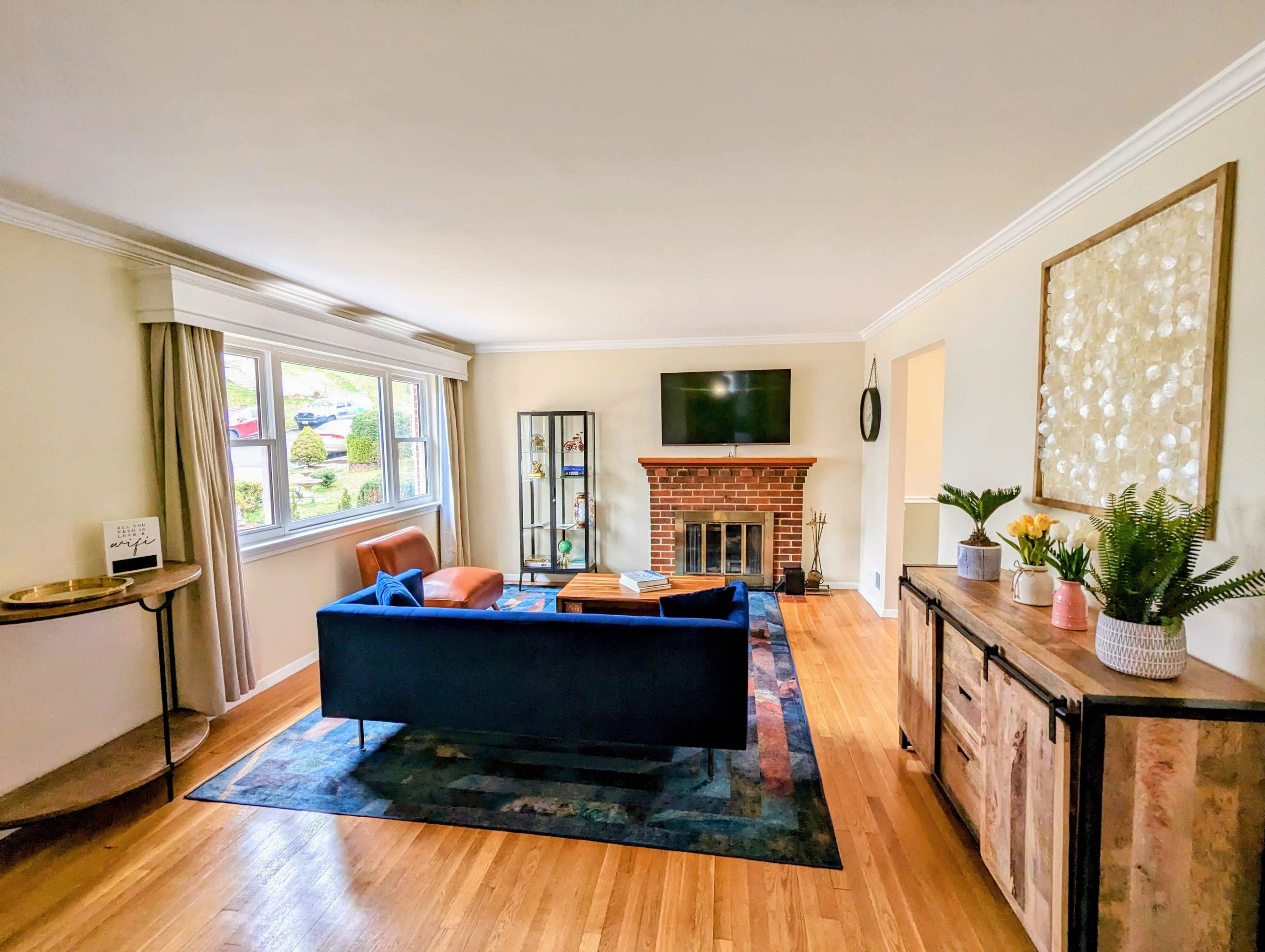 The living room features a blue sofa, a brick fireplace, a TV mounted on the wall, and a wooden sideboard, with large windows allowing natural light.