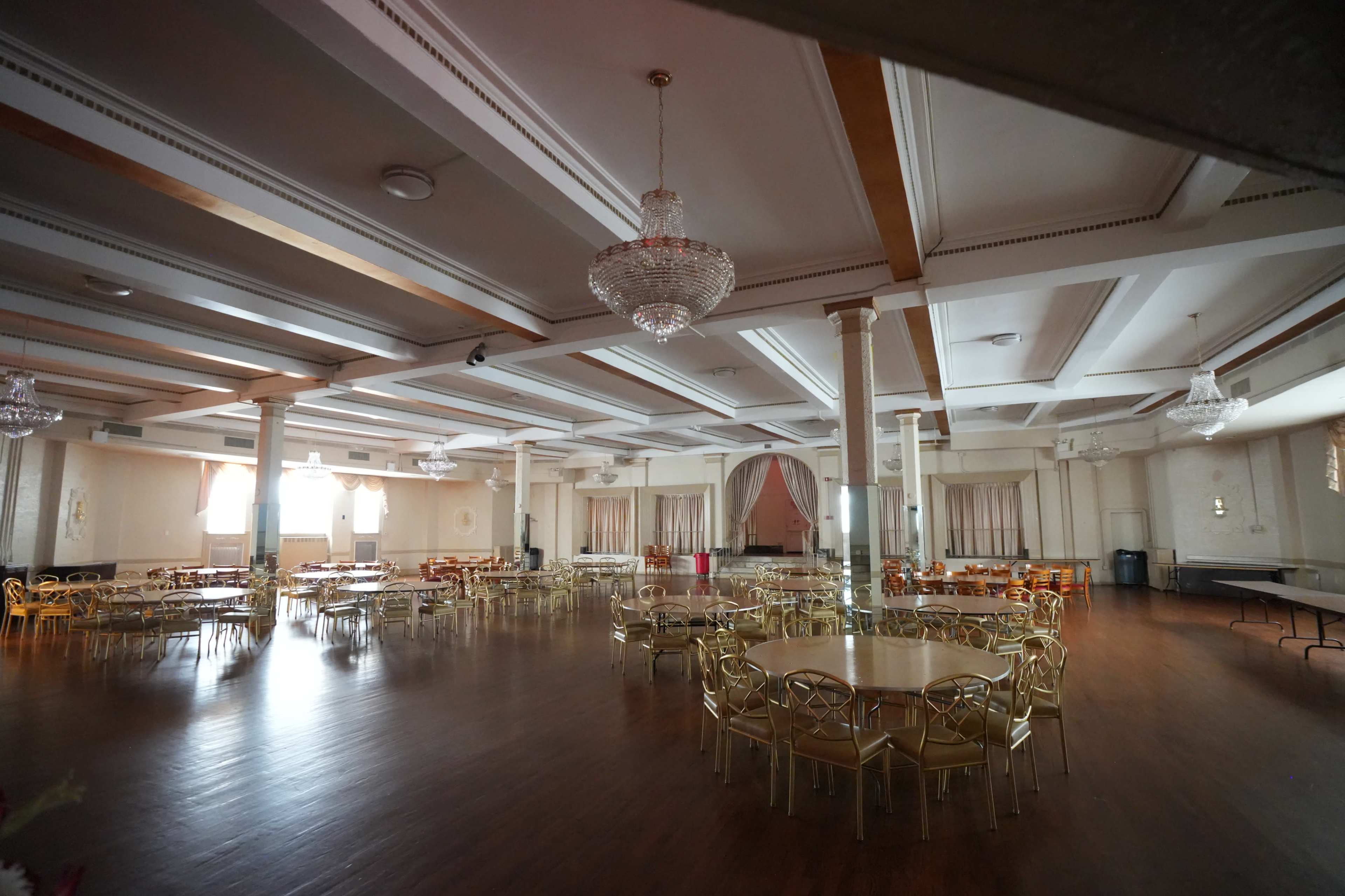 The image shows a spacious and empty banquet hall with round tables and gold-toned chairs arranged across a polished wooden floor, illuminated by overhead lights and adorned with chandeliers.