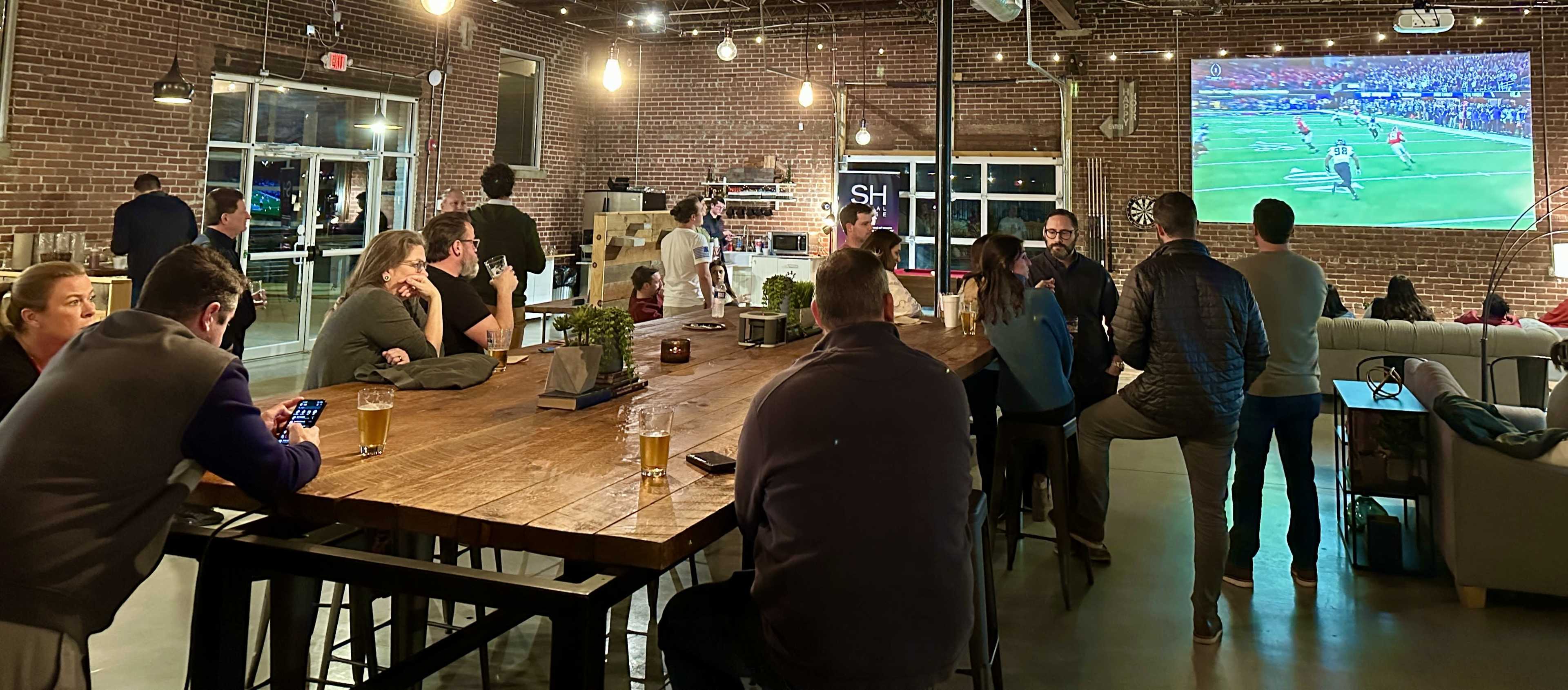 A group of people sits at a long wooden bar while watching a sports game projected on a wall in a casual indoor setting.
