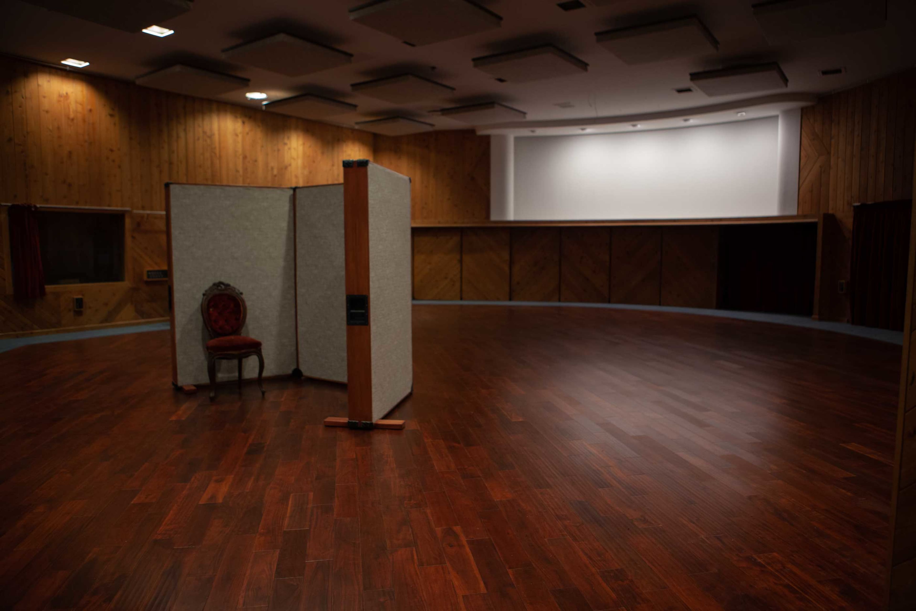 An empty room featuring a wooden floor, a large screen at one end, and a folding partition with a lone chair beside it.