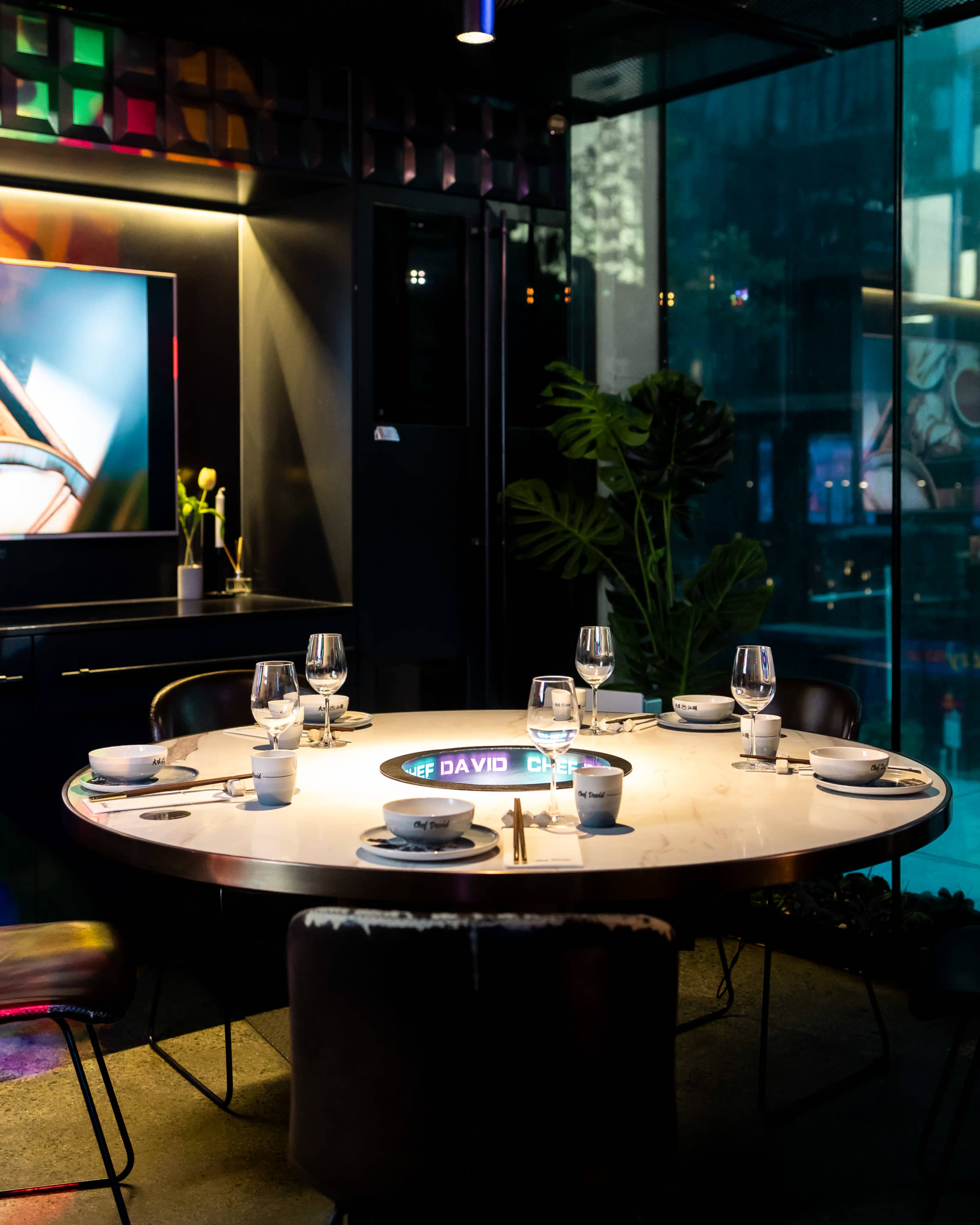 A round dining table is set with plates and glasses in a modern restaurant with a colorful backdrop and glass walls.
