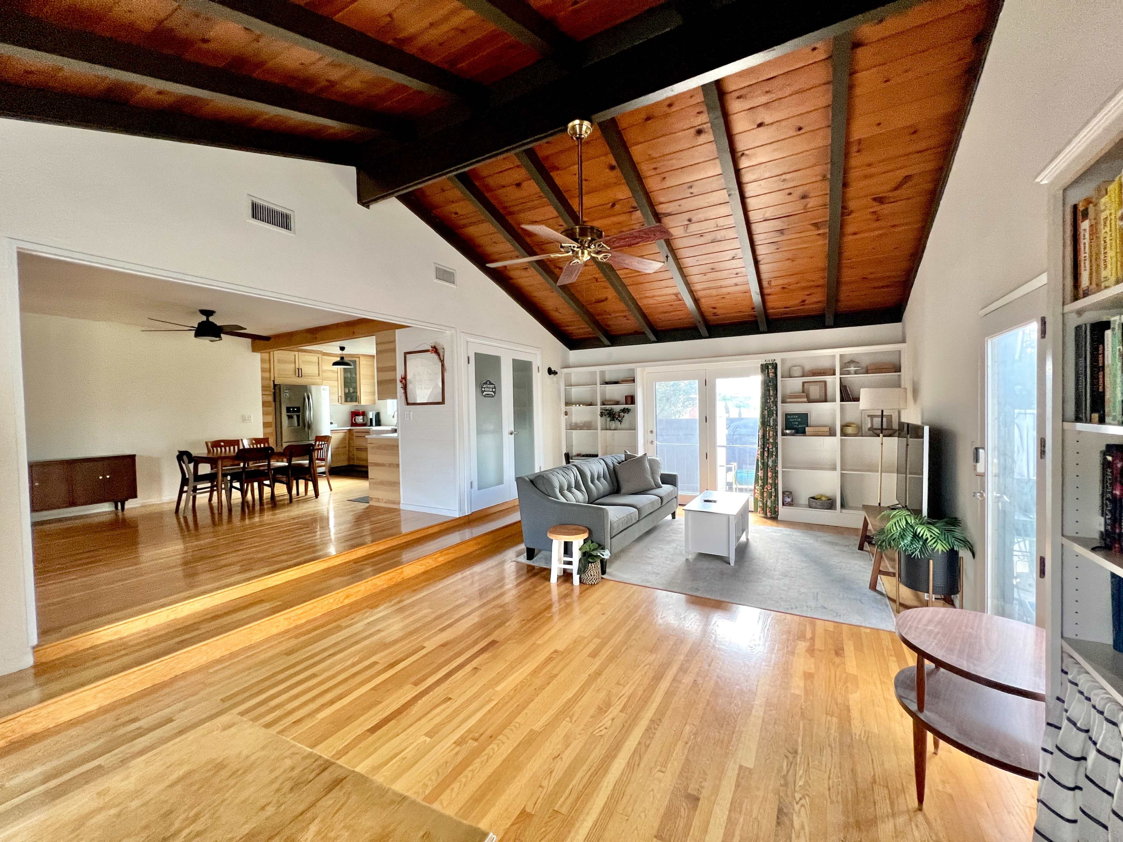 A spacious living area features a vaulted wooden ceiling, hardwood floors, and an open layout with a seating area and shelving.