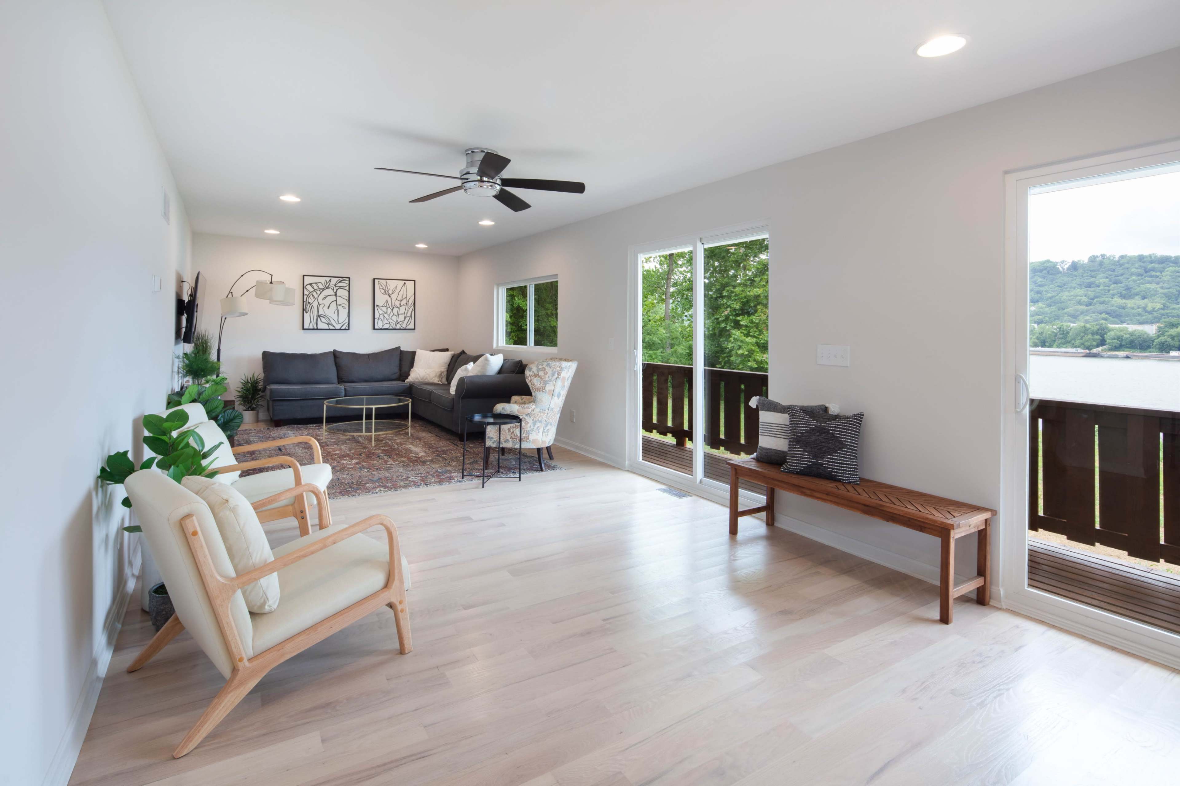 The room features a modern living area with a sectional sofa, a ceiling fan, large windows, and a wooden bench, overlooking a scenic view.