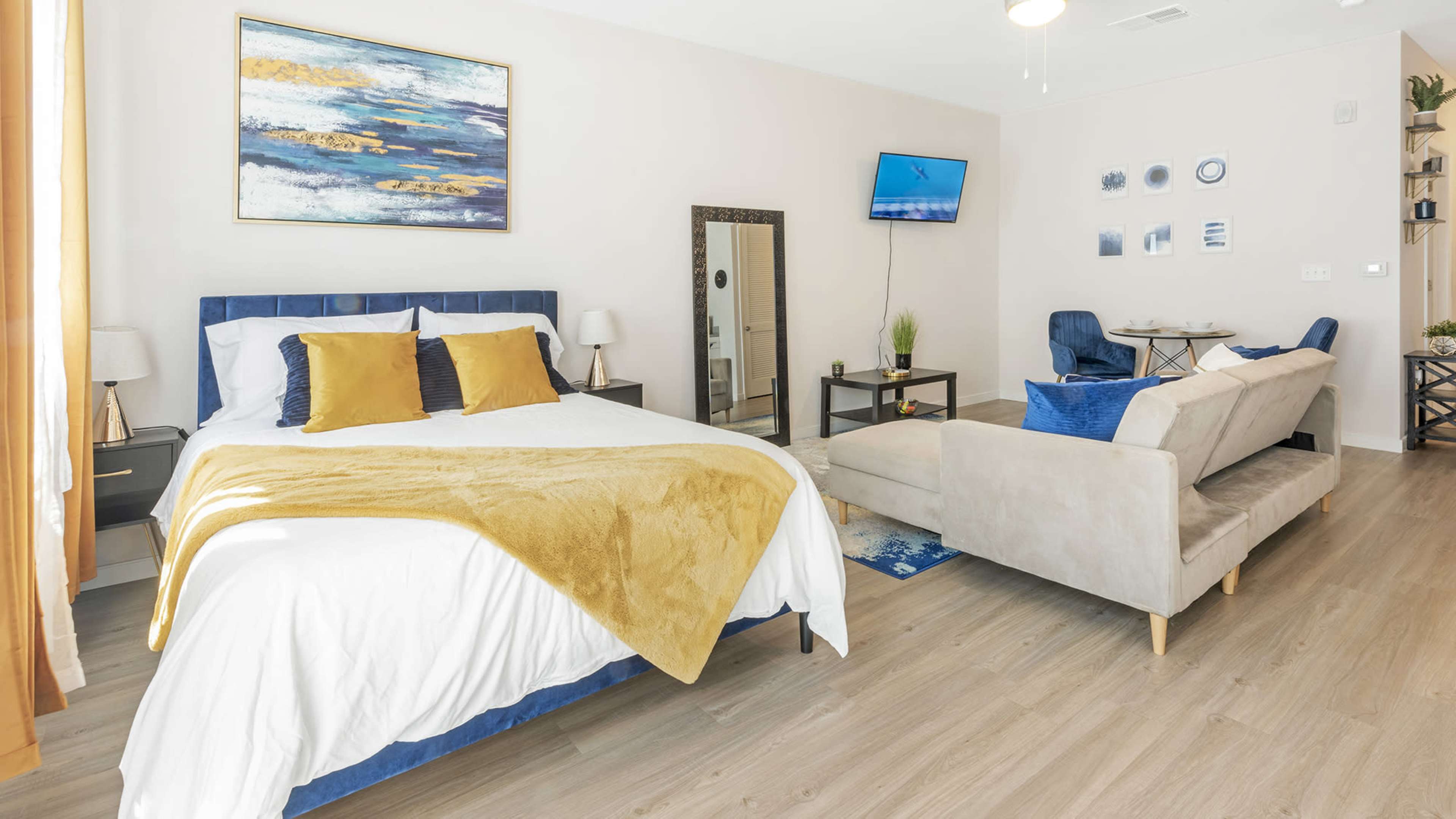 The image shows a well-decorated bedroom with a blue bed, a beige couch, a mirror, and a television mounted on the wall.