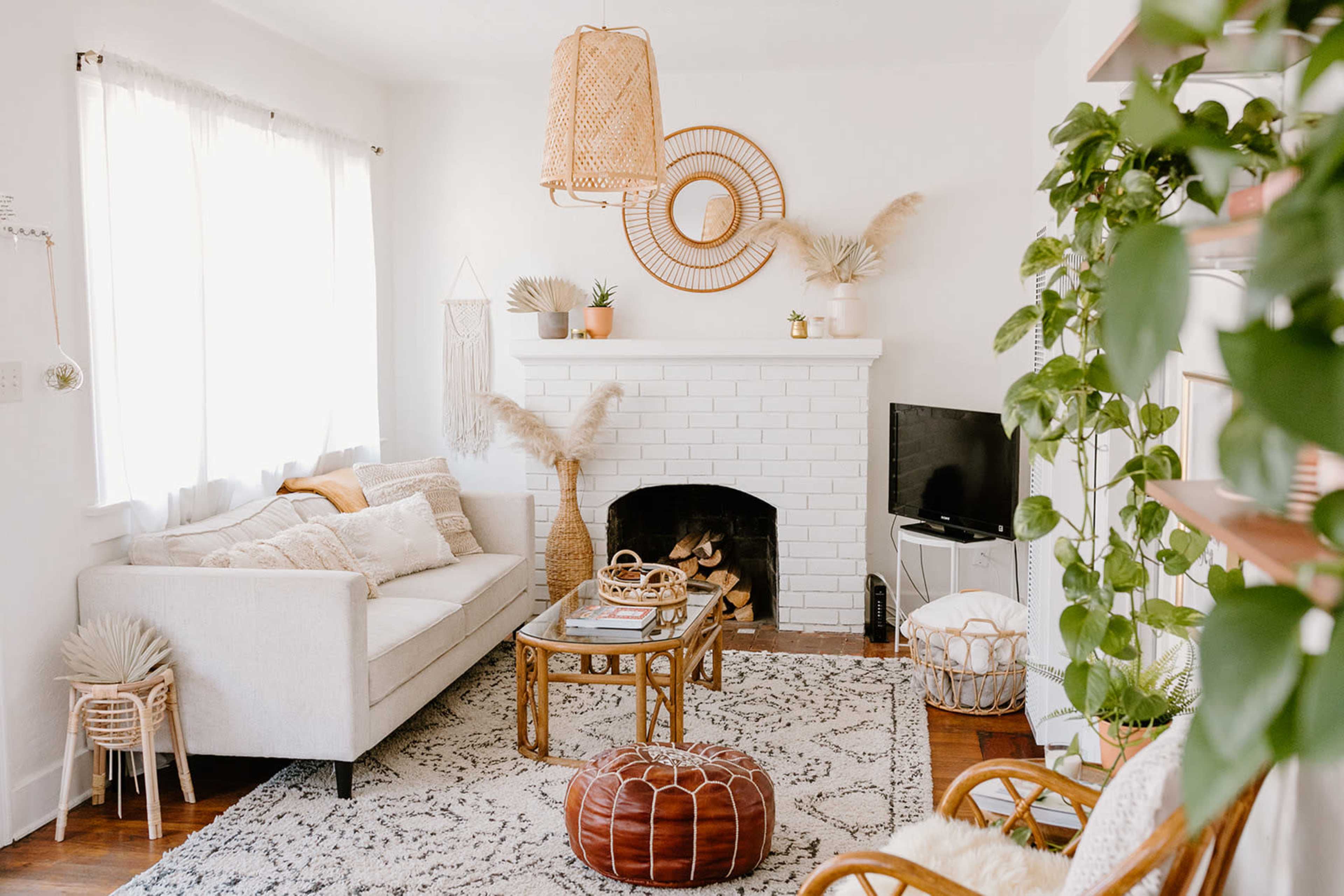 A cozy living room features a white brick fireplace, a light-colored sofa, a round mirror above the mantel, and a variety of plants throughout the space.