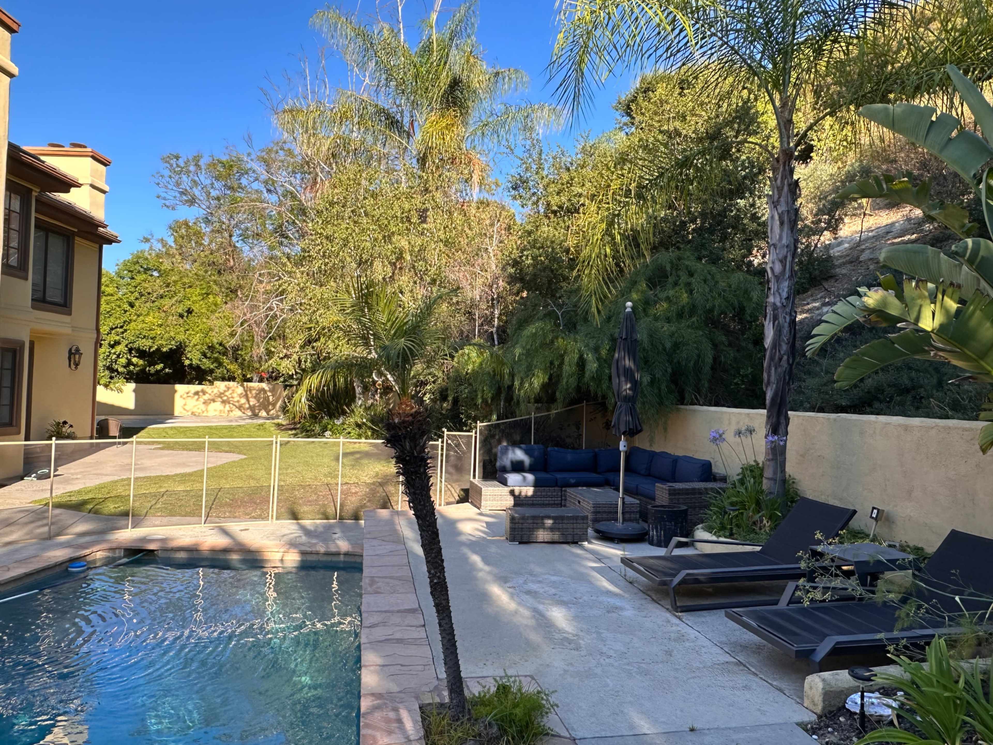 A backyard with a swimming pool, lounge chairs, a patio area with seating, and a grassy space surrounded by trees and shrubs.