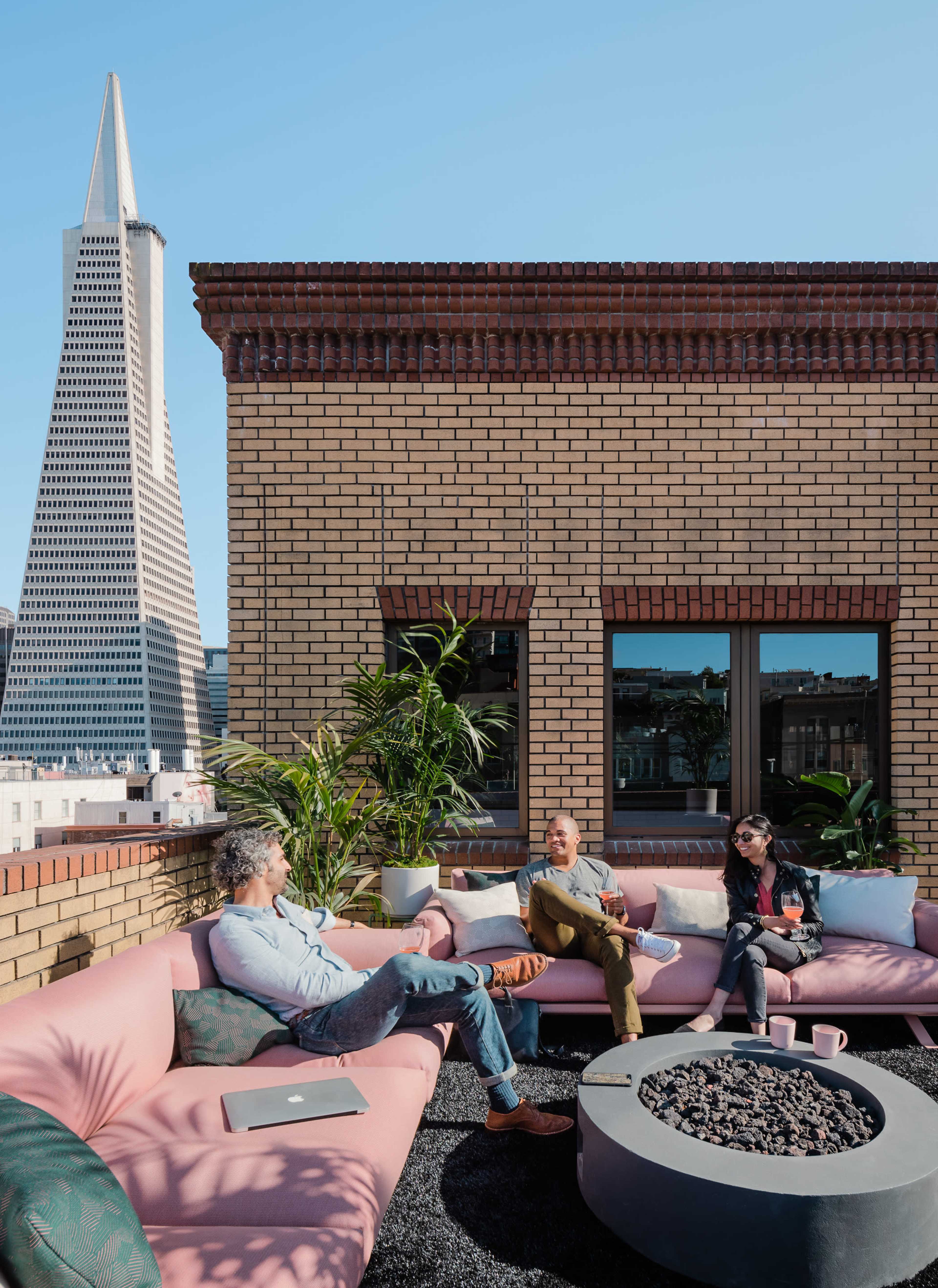 Three people are seated on a pink outdoor sofa on a rooftop, with a modern skyscraper in the background.