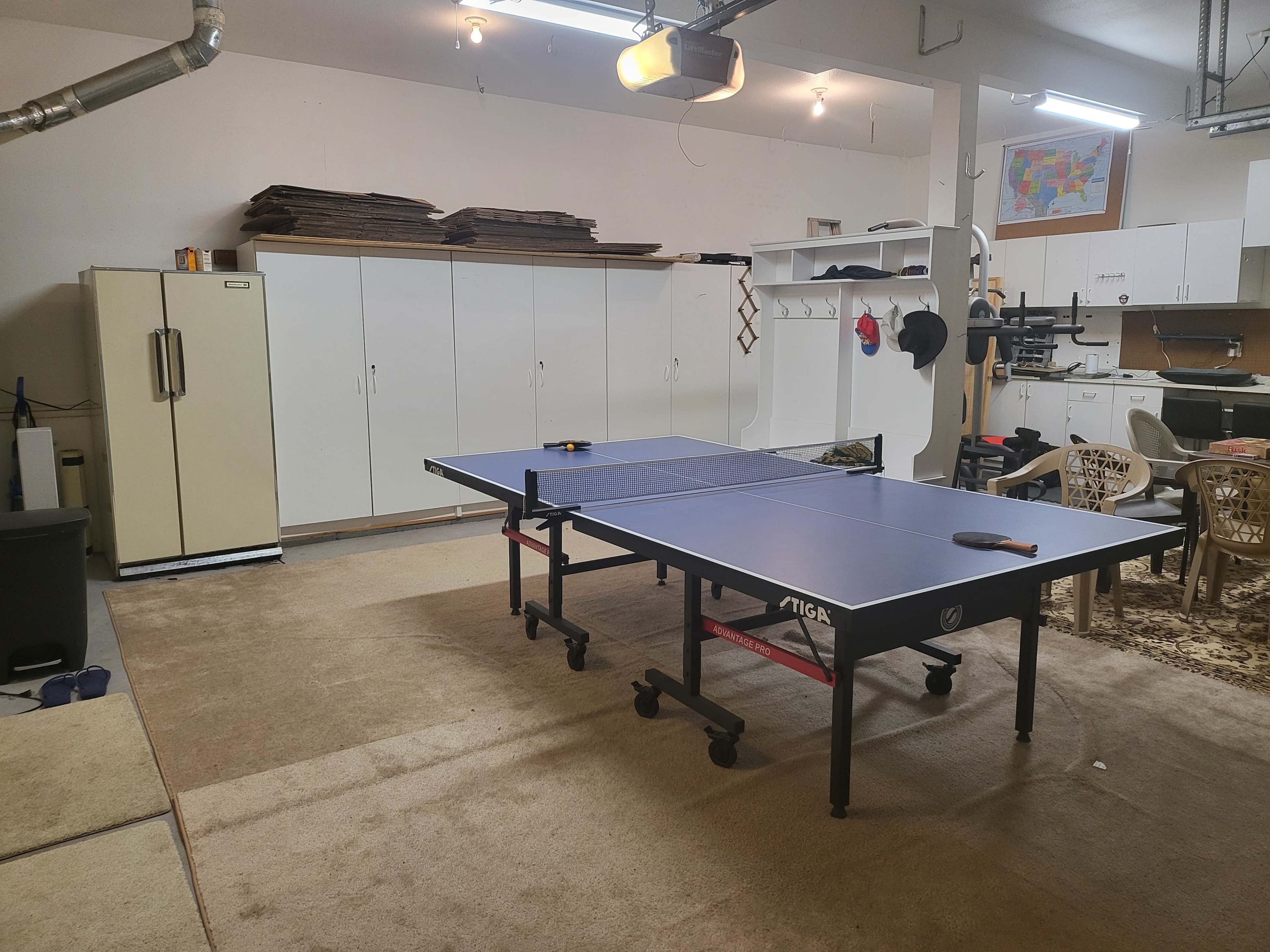 A garage-style room with a ping pong table in the center, surrounded by cabinets and a kitchenette area.