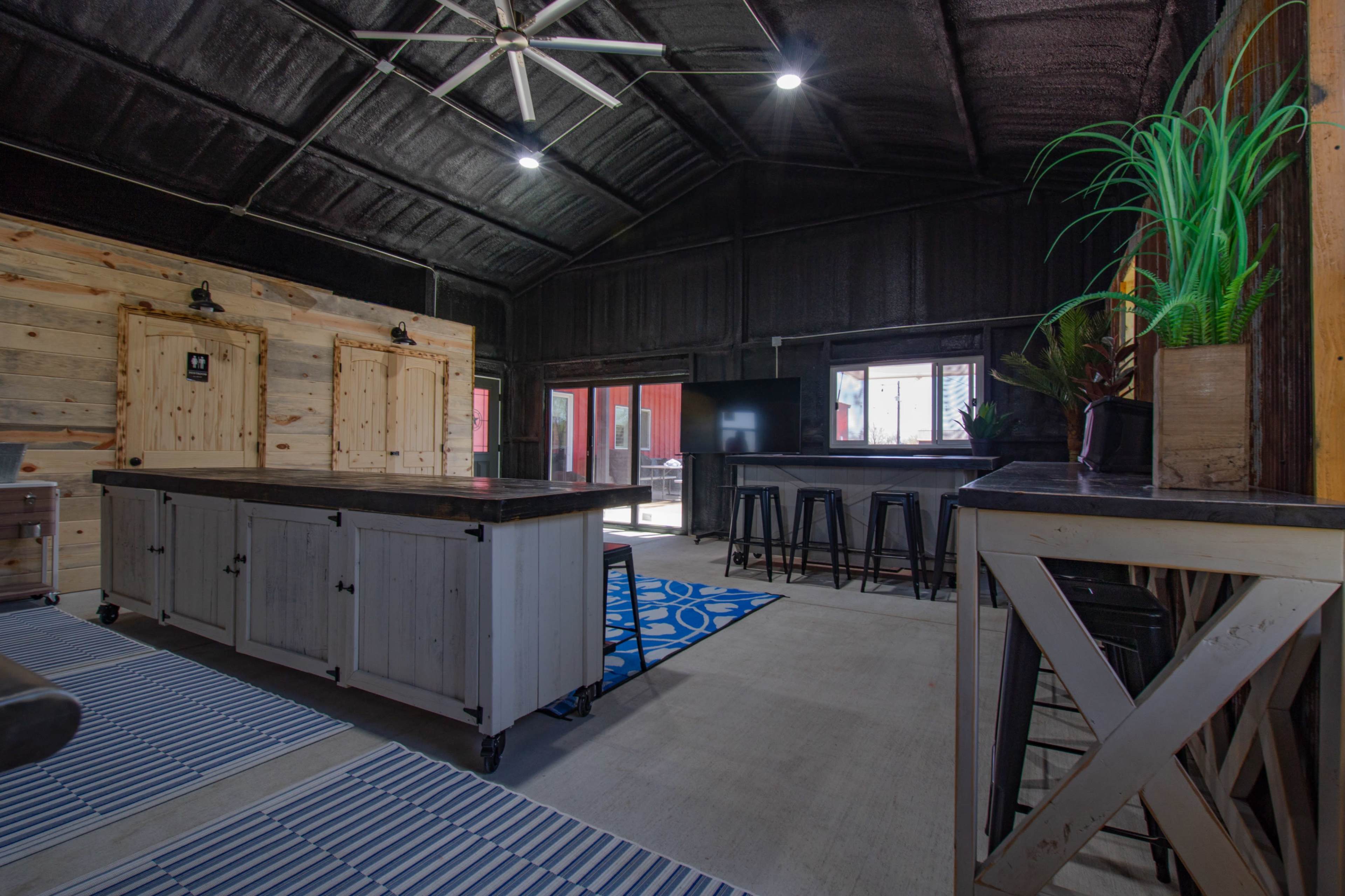The image shows a spacious interior of a modern gathering space with a large wooden table, bar stools, and a ceiling fan, featuring wooden walls and large windows.