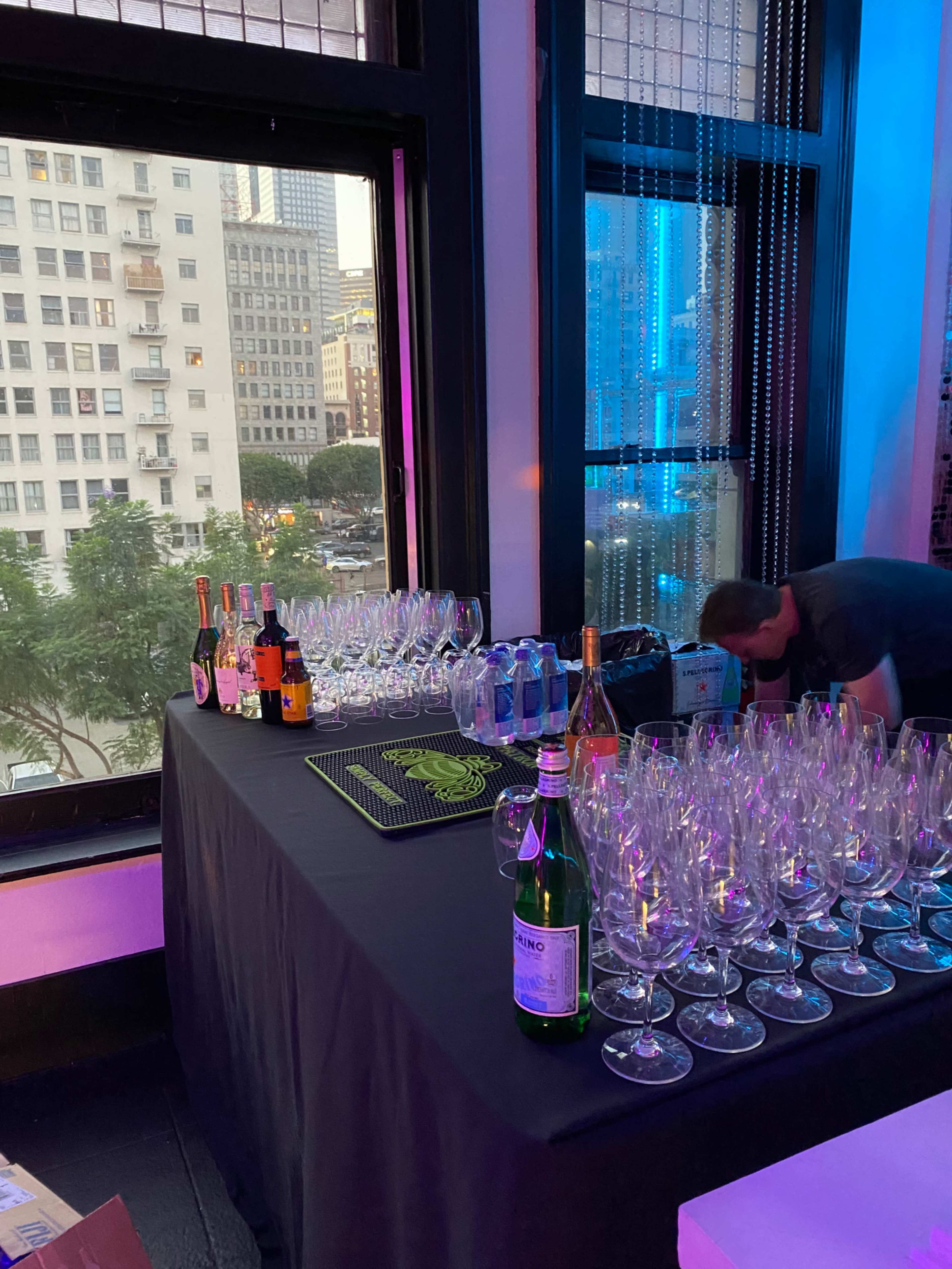 A table is set with various glasses, bottles of wine and champagne, and a bartender preparing in a brightly lit room with large windows overlooking a cityscape.