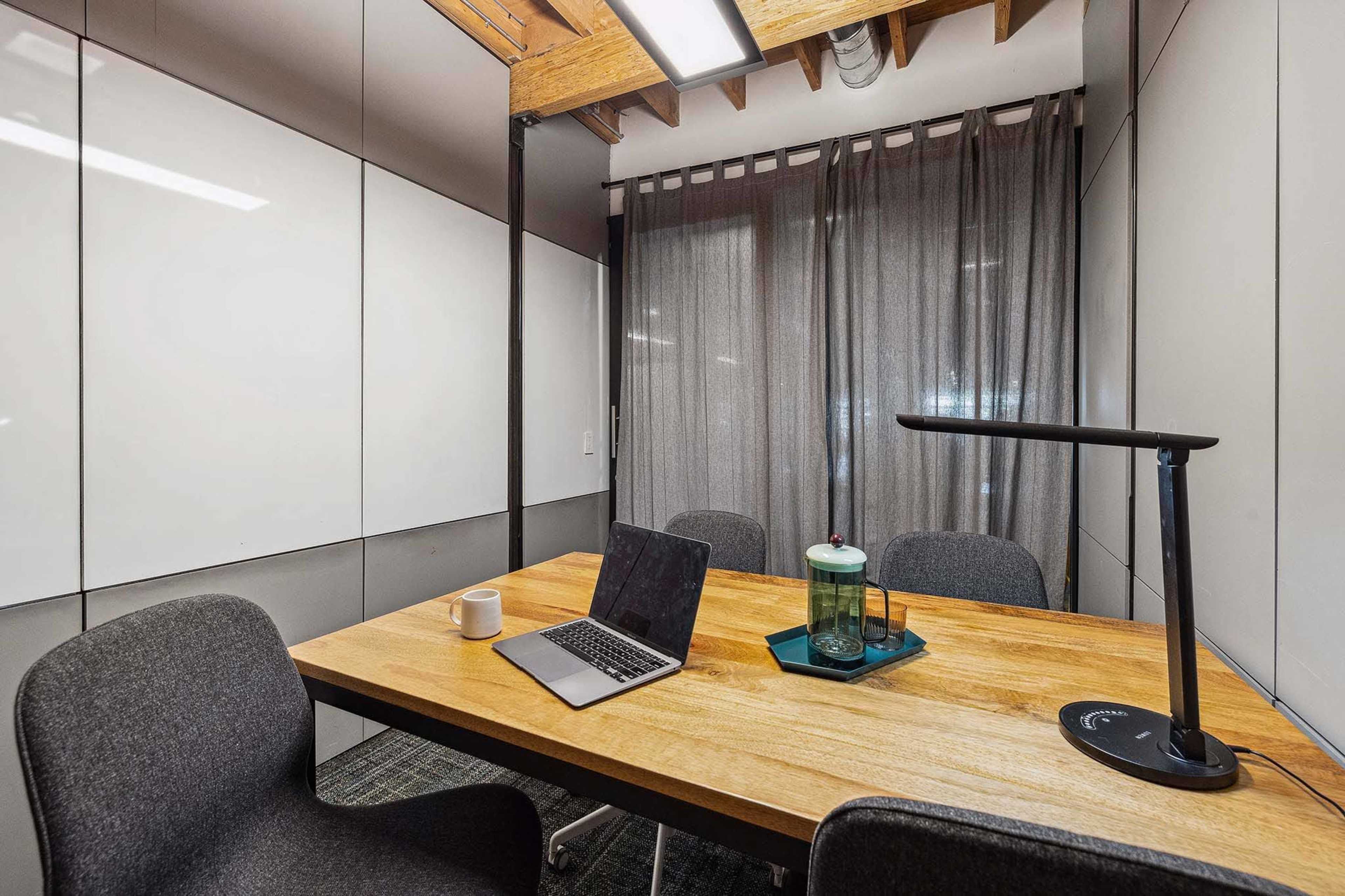 A modern meeting room features a wooden table with a laptop, a cup, and a green glass container, surrounded by three chairs and gray curtains.