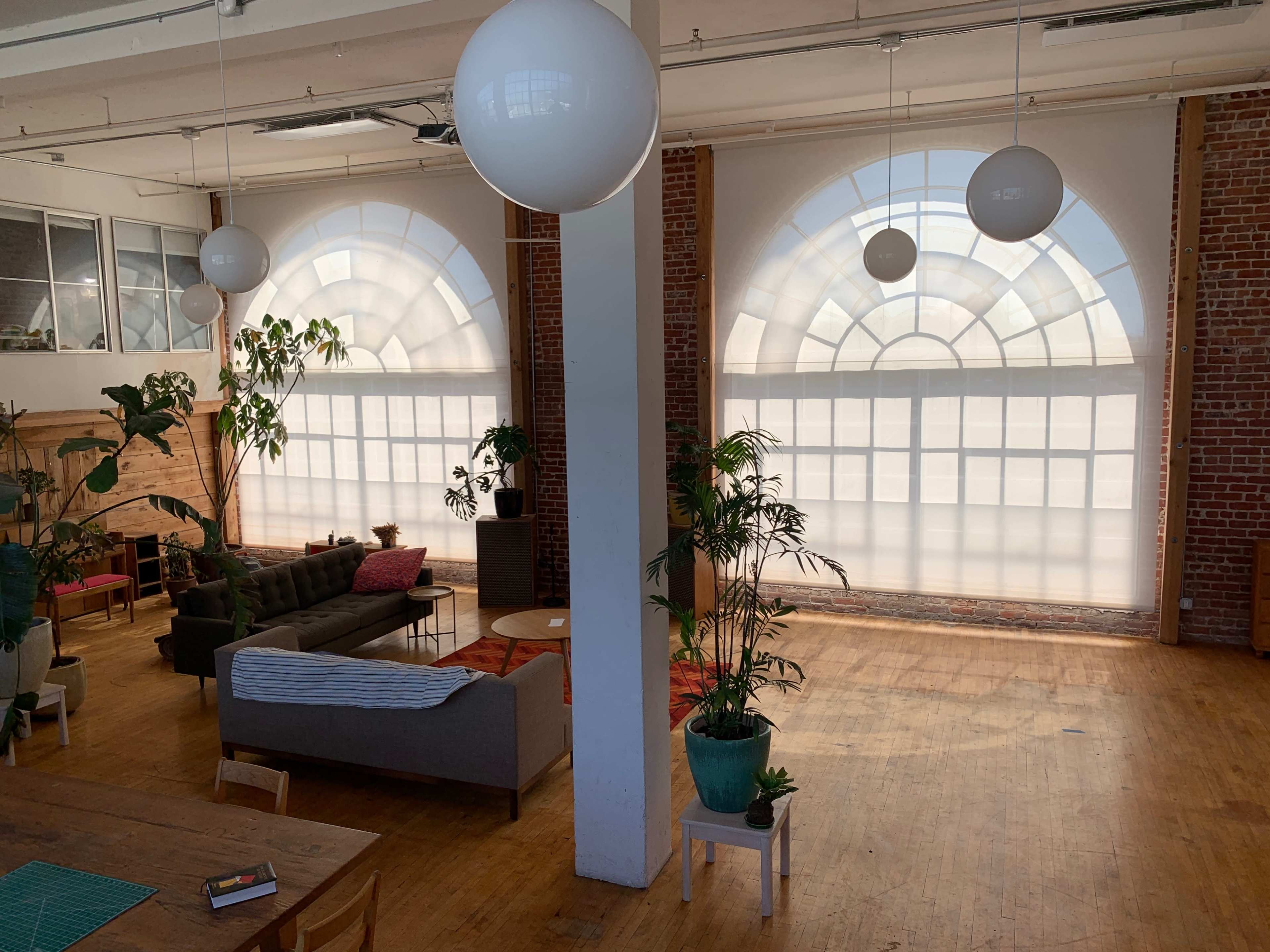 The room features large arched windows, a cozy seating area with a sofa and coffee table, and several indoor plants on wooden floors.