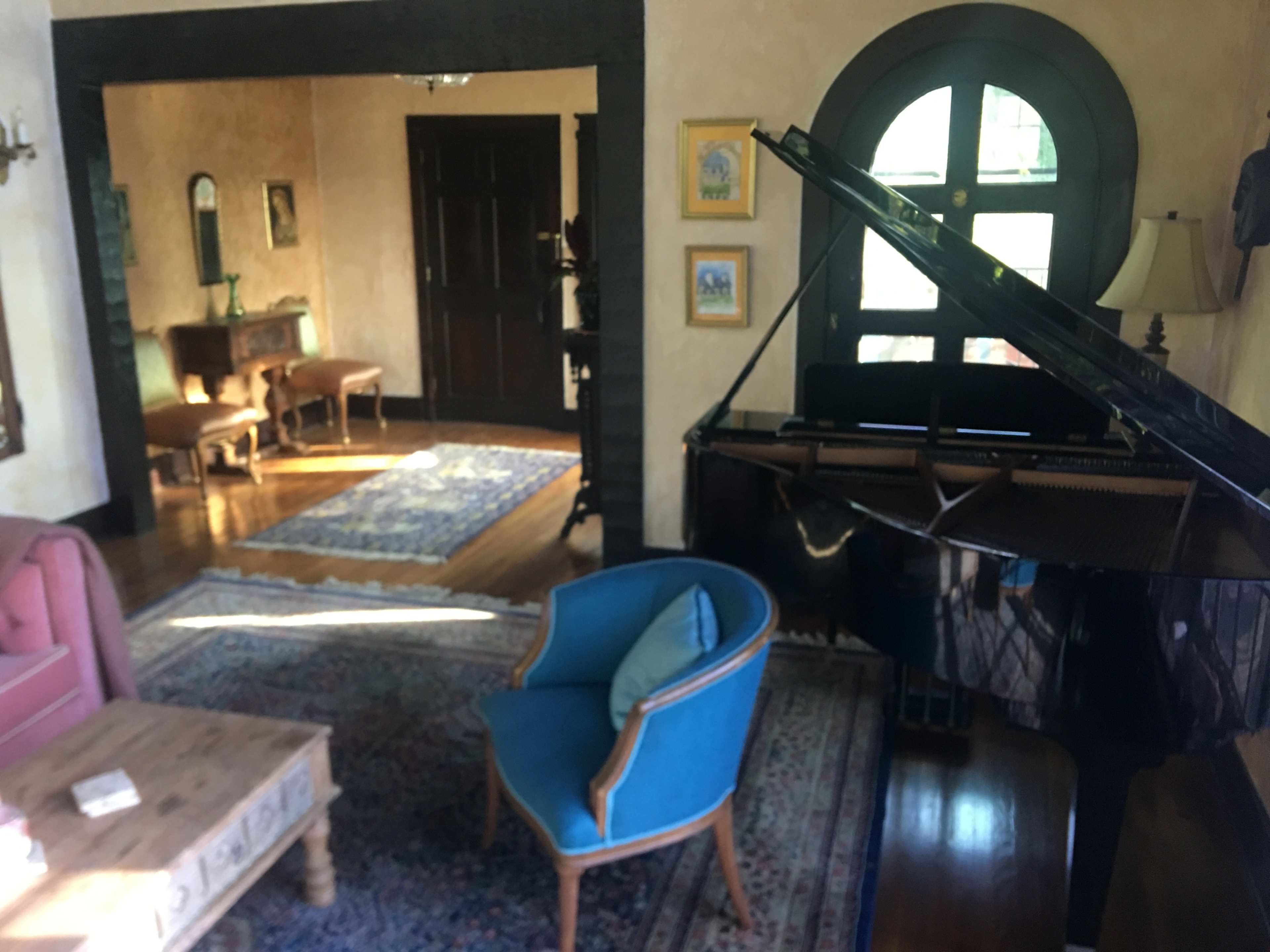 A grand piano sits next to a blue armchair in a well-furnished living room with wooden floors and large windows.