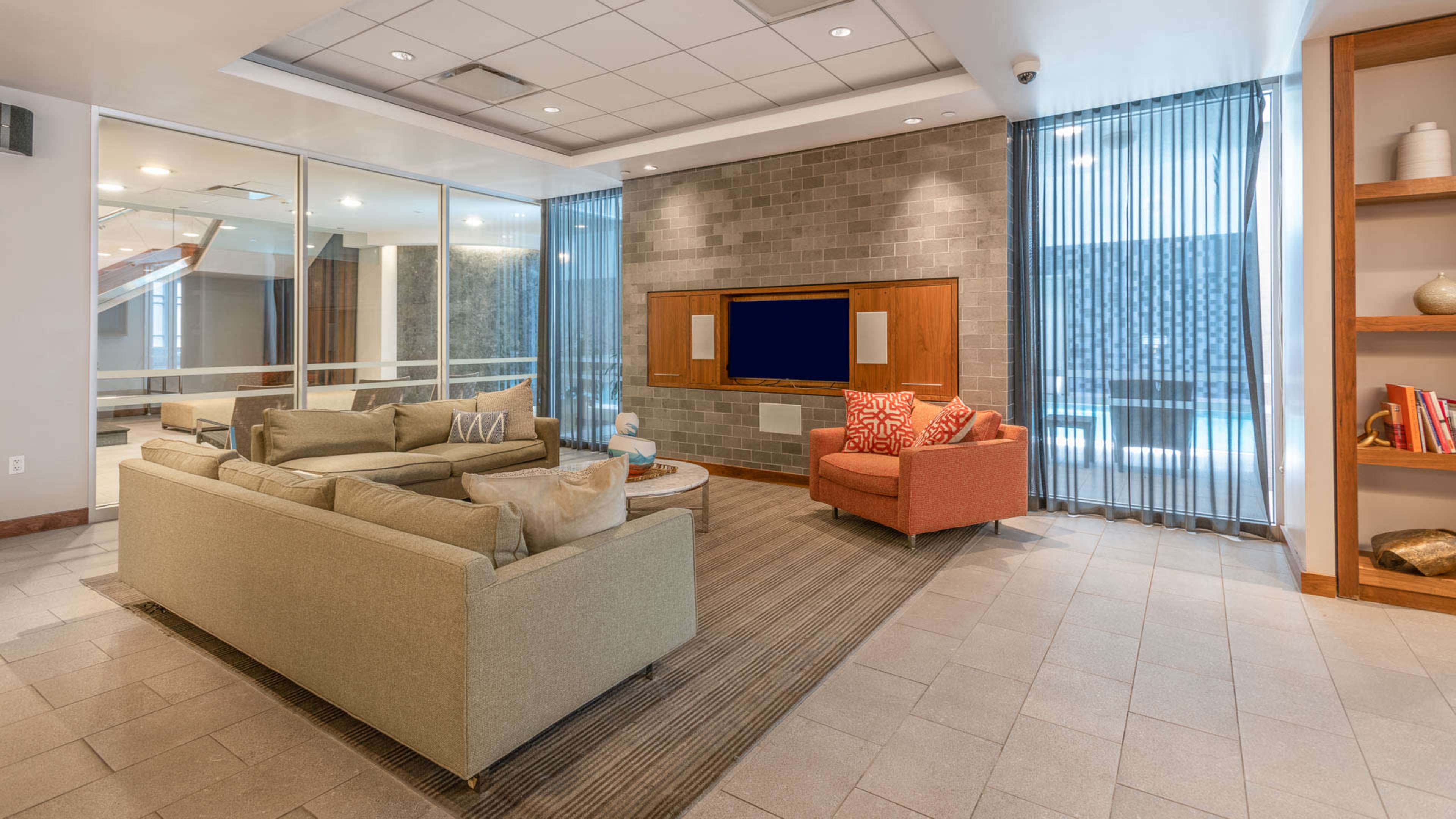 The image shows a modern lounge area featuring a sofa, an armchair, a coffee table, and a TV mounted on a textured wall, with large windows draped in sheer curtains.