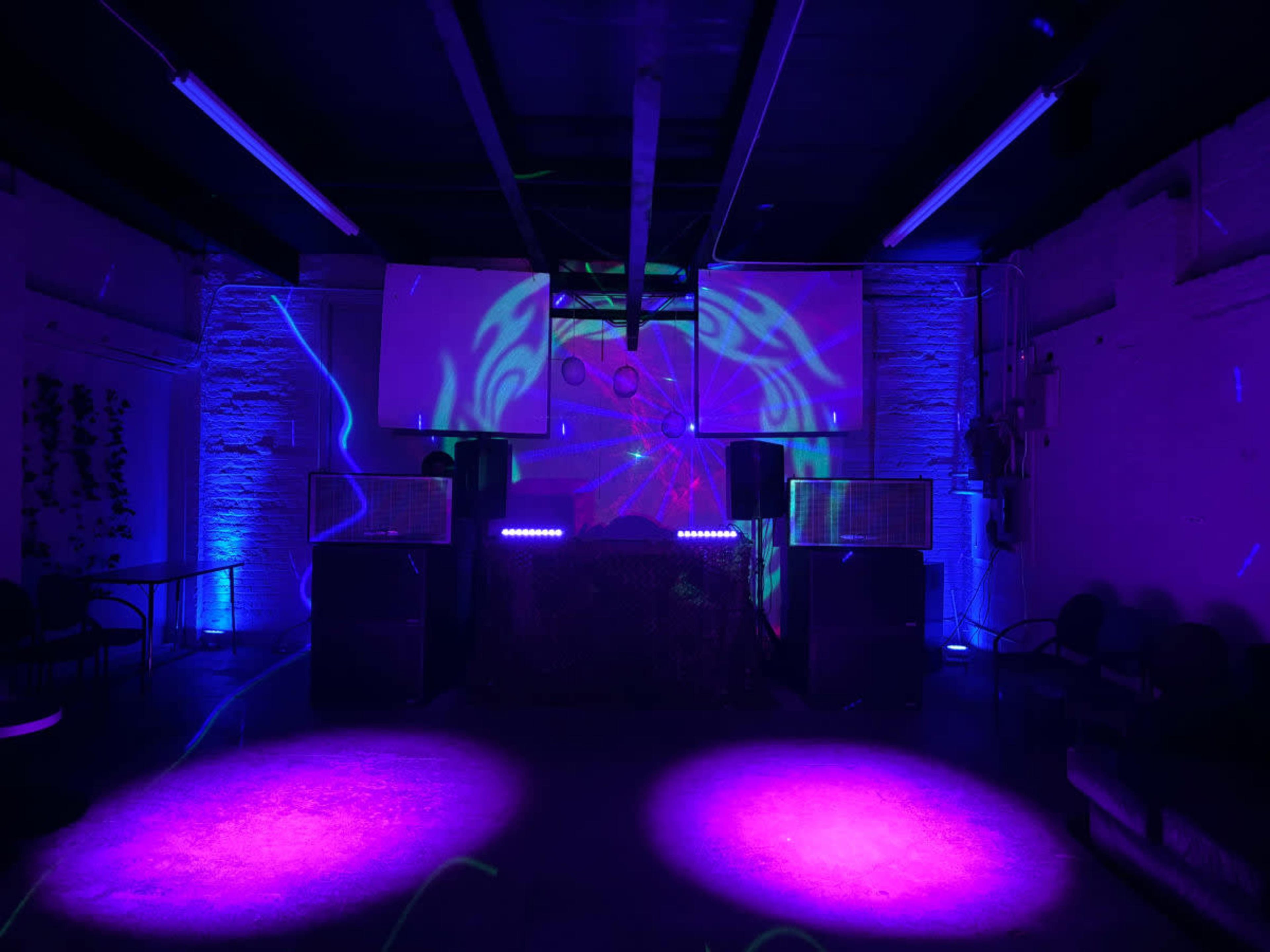 A dimly lit indoor venue with a DJ setup featuring large speakers, colorful lights, and a projection on a wall.