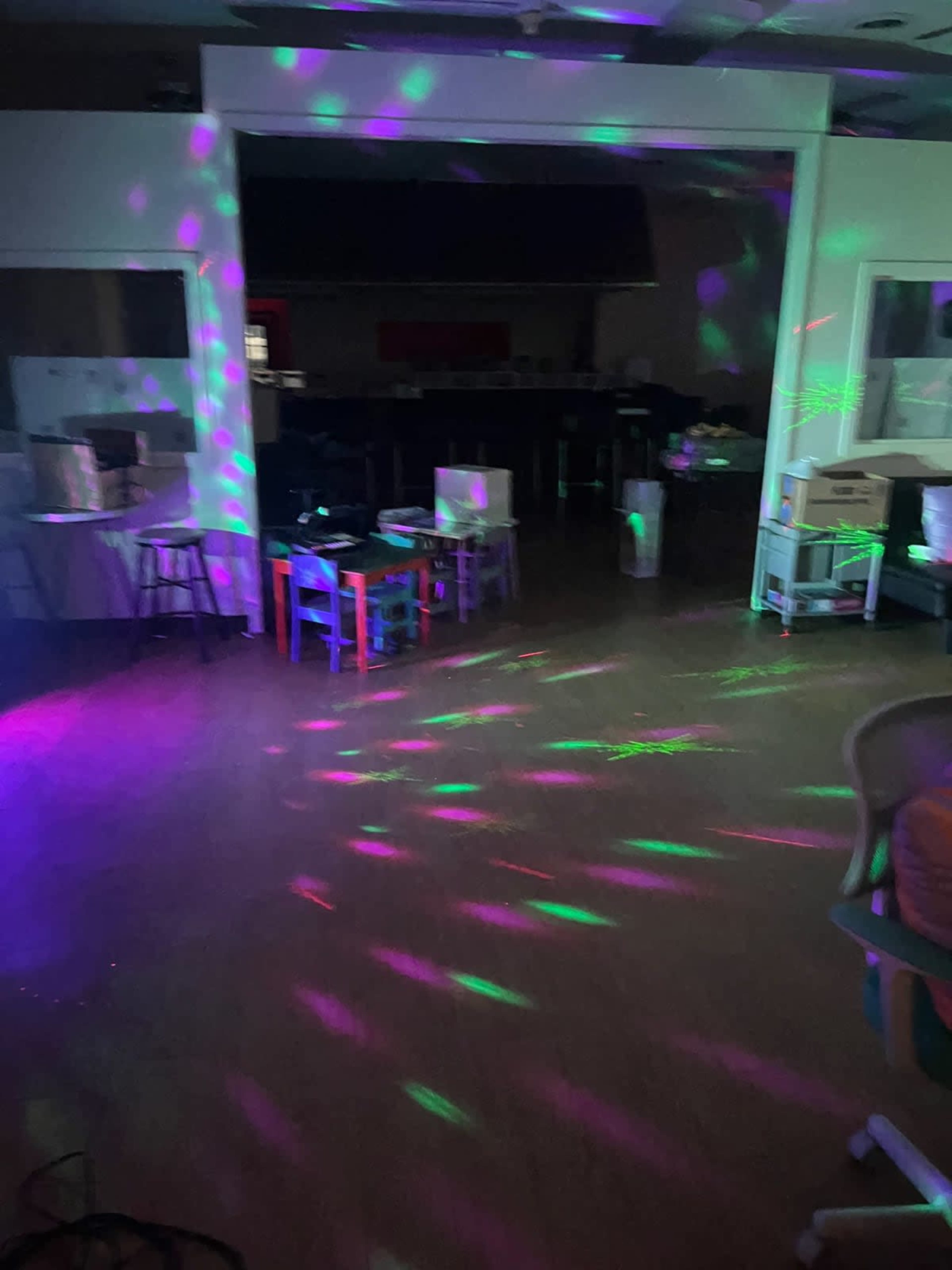 The image shows a dimly lit room with colorful light patterns projected onto the floor and walls, and several tables and chairs scattered throughout.