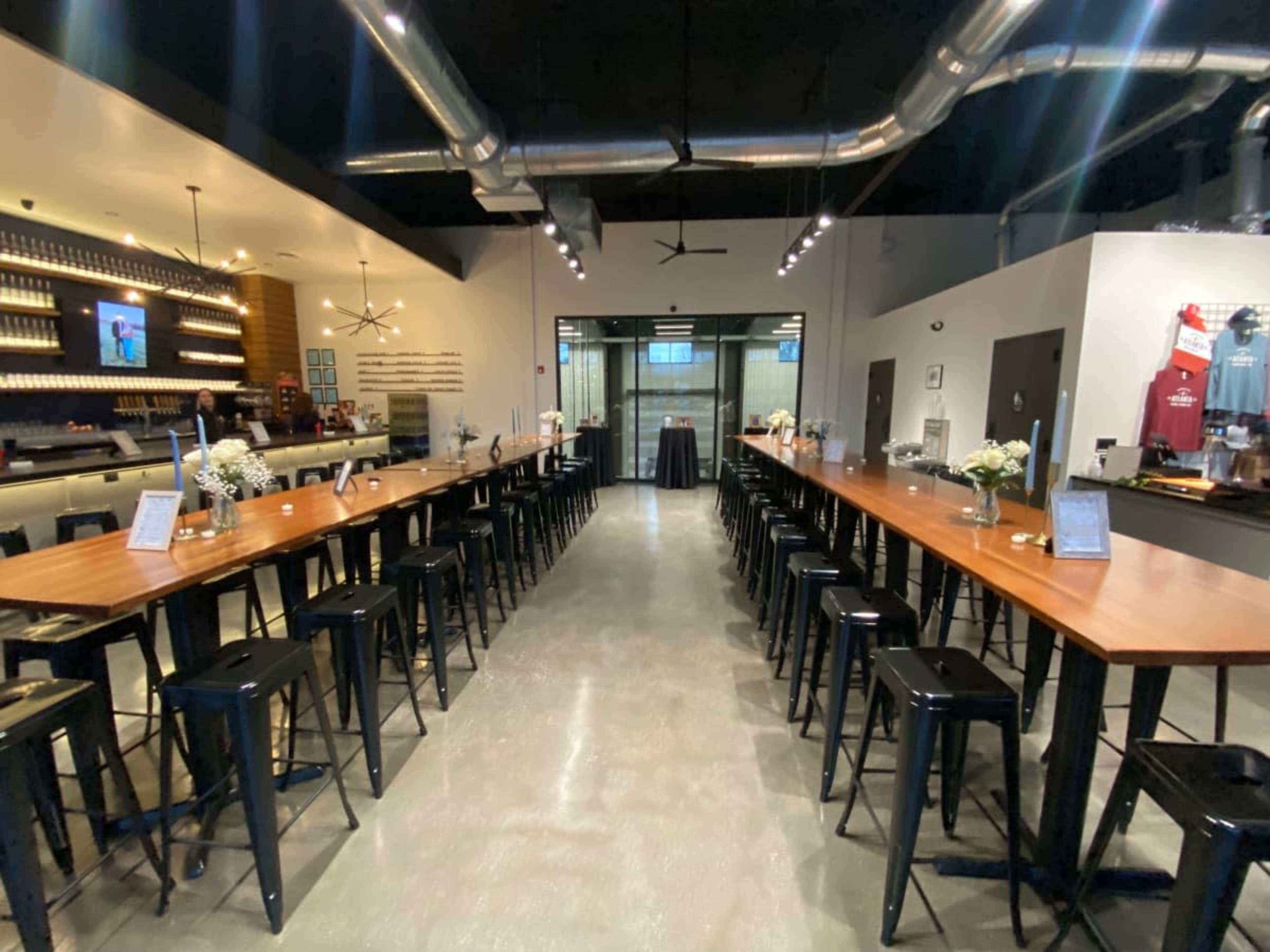 A modern bar interior with a long, wooden communal table flanked by black metal stools, and a well-lit service area in the background.