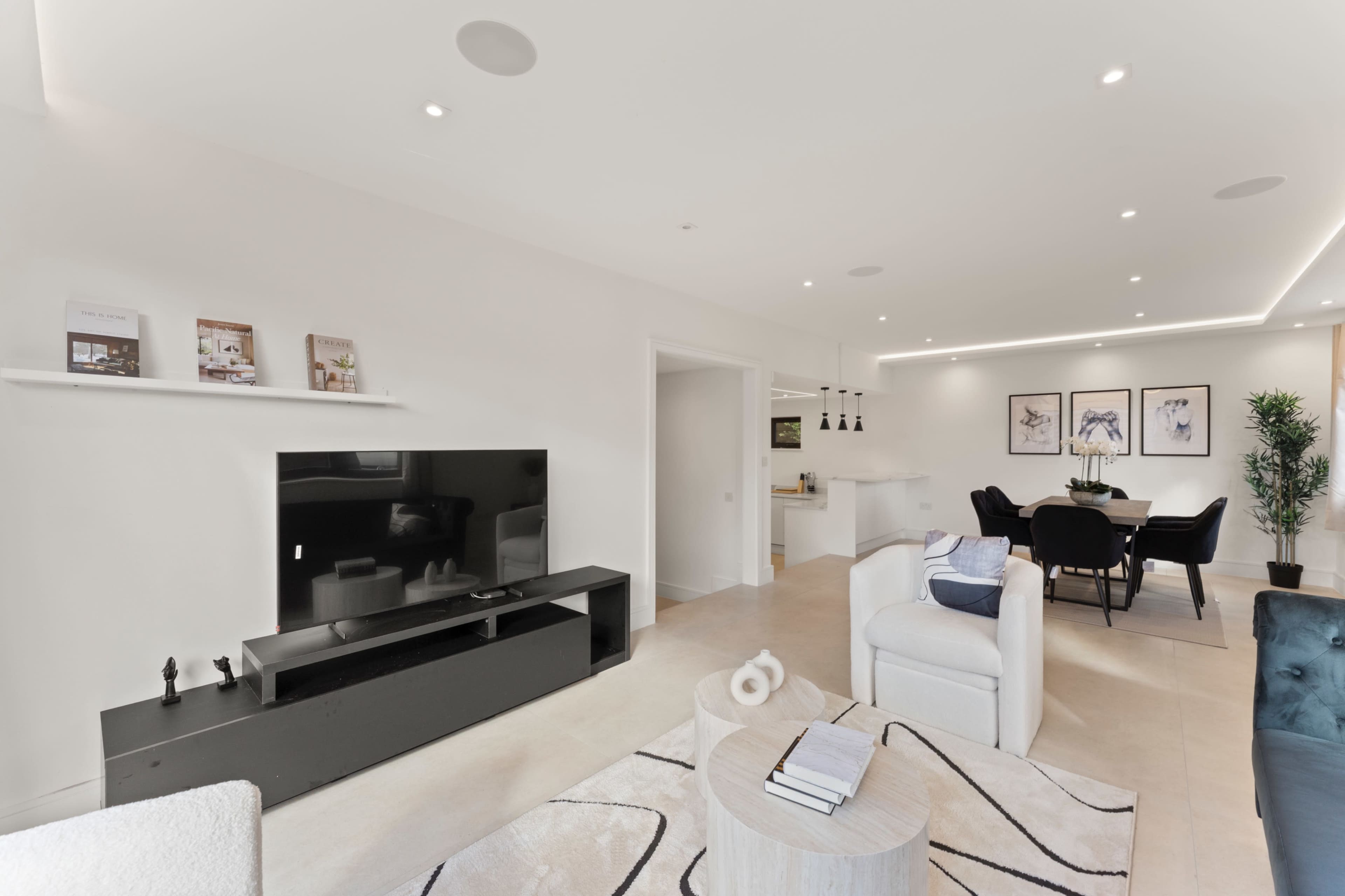 The image shows a modern living room with a black television unit, light-colored walls, and a dining area featuring a round table and black chairs.