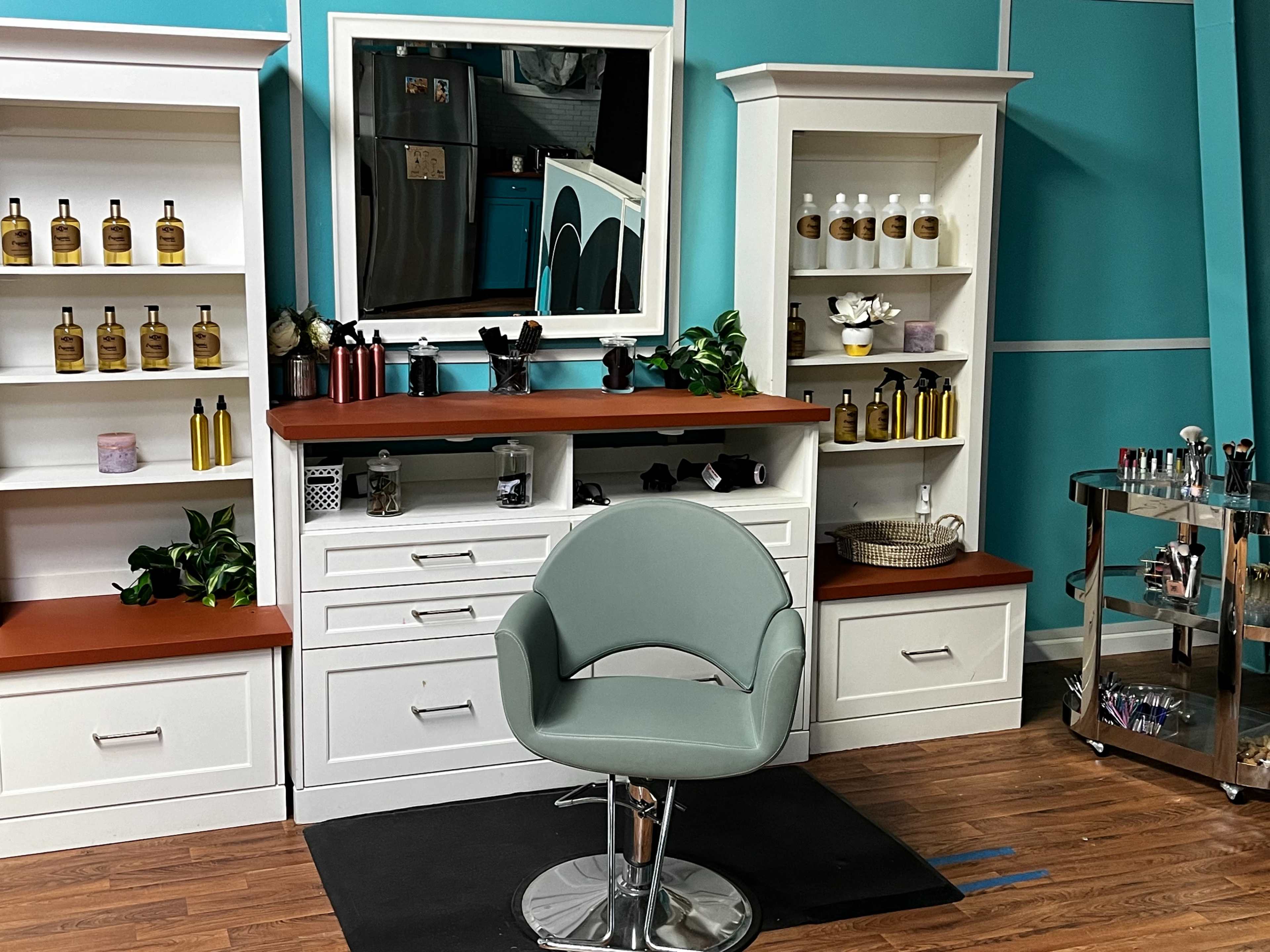 The image shows a hair salon interior with a mirrored vanity, organized shelves containing beauty products, and a styling chair positioned in front of the mirror.
