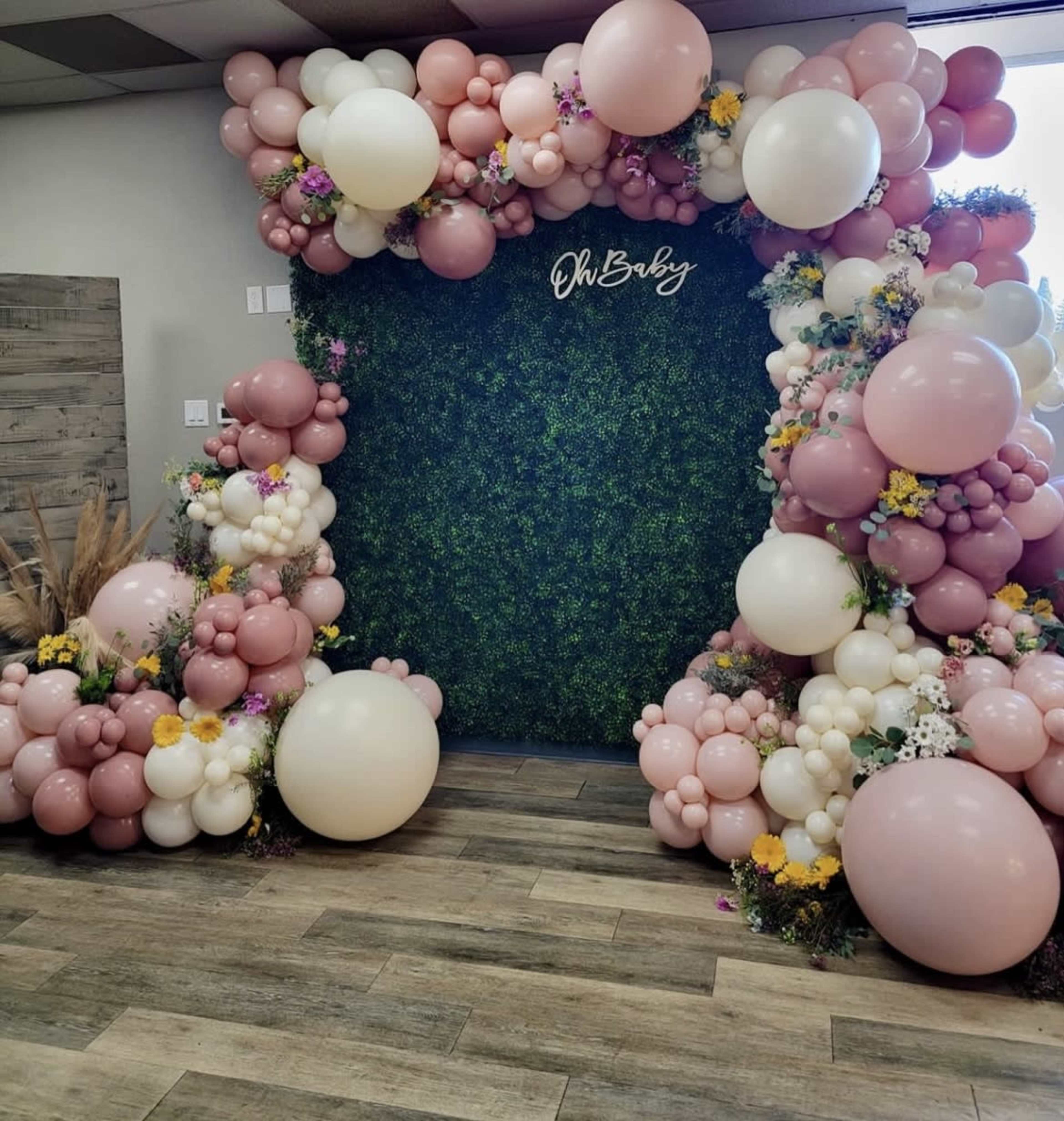 The image shows a decorative backdrop made of greenery, adorned with arranged balloons in shades of pink, white, and cream, with some floral elements surrounding the structure.