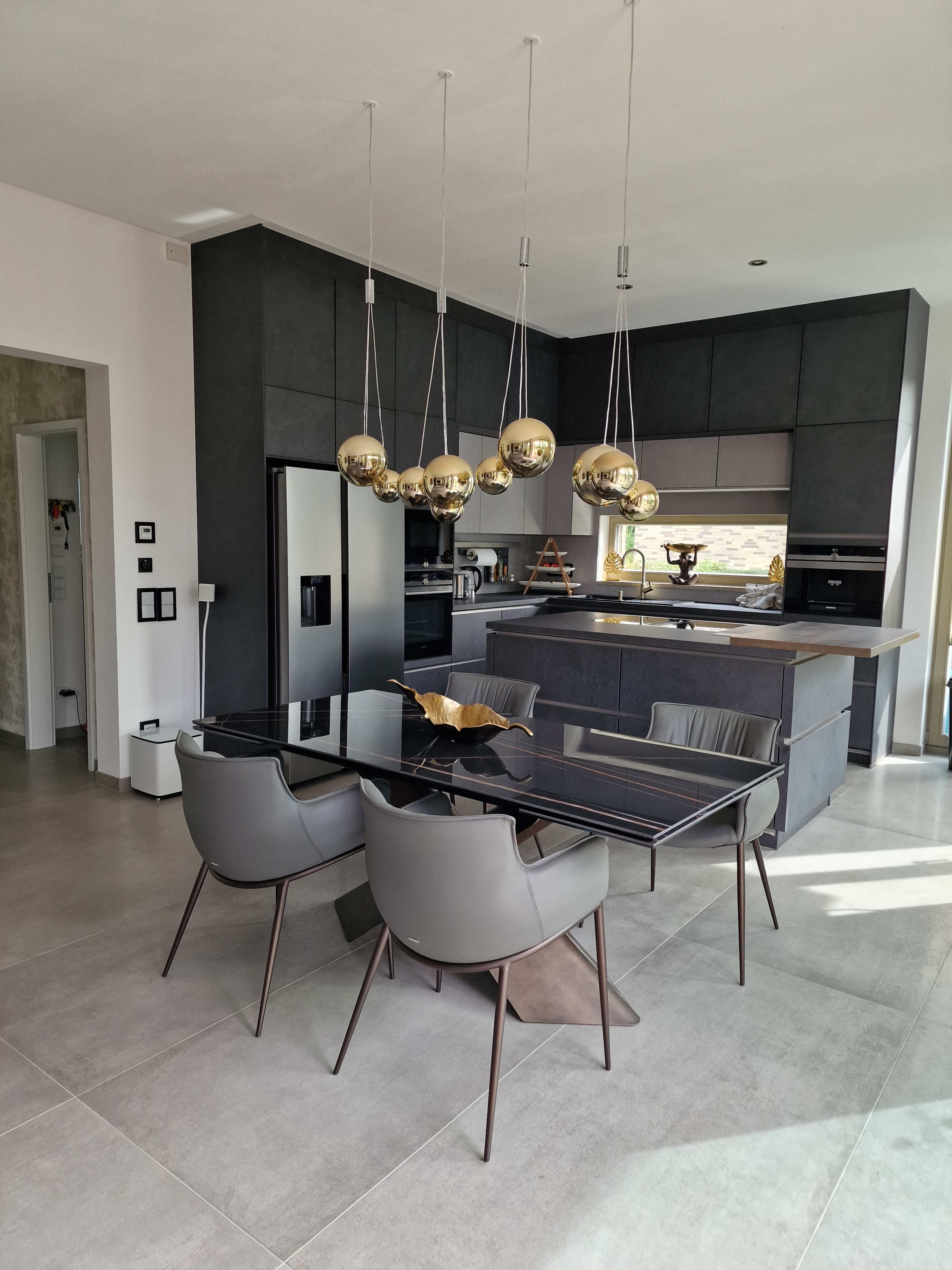 A modern kitchen features a glass-top dining table with four chairs and pendant lights above, complemented by dark cabinetry and stainless steel appliances.
