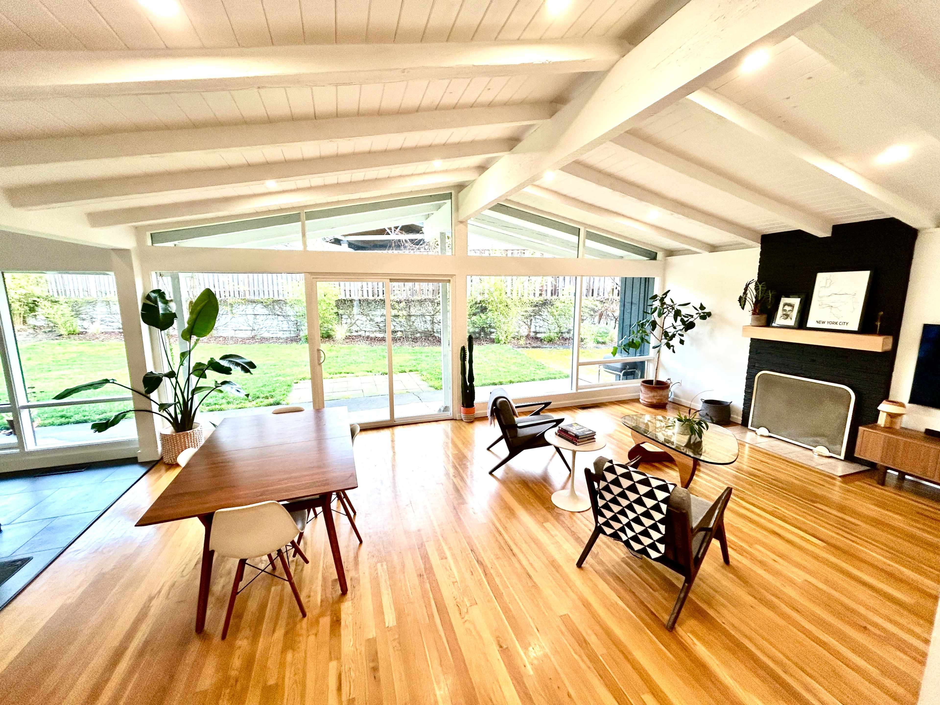 A bright interior living space featuring a wooden dining table, modern chairs, large windows, and a fireplace.