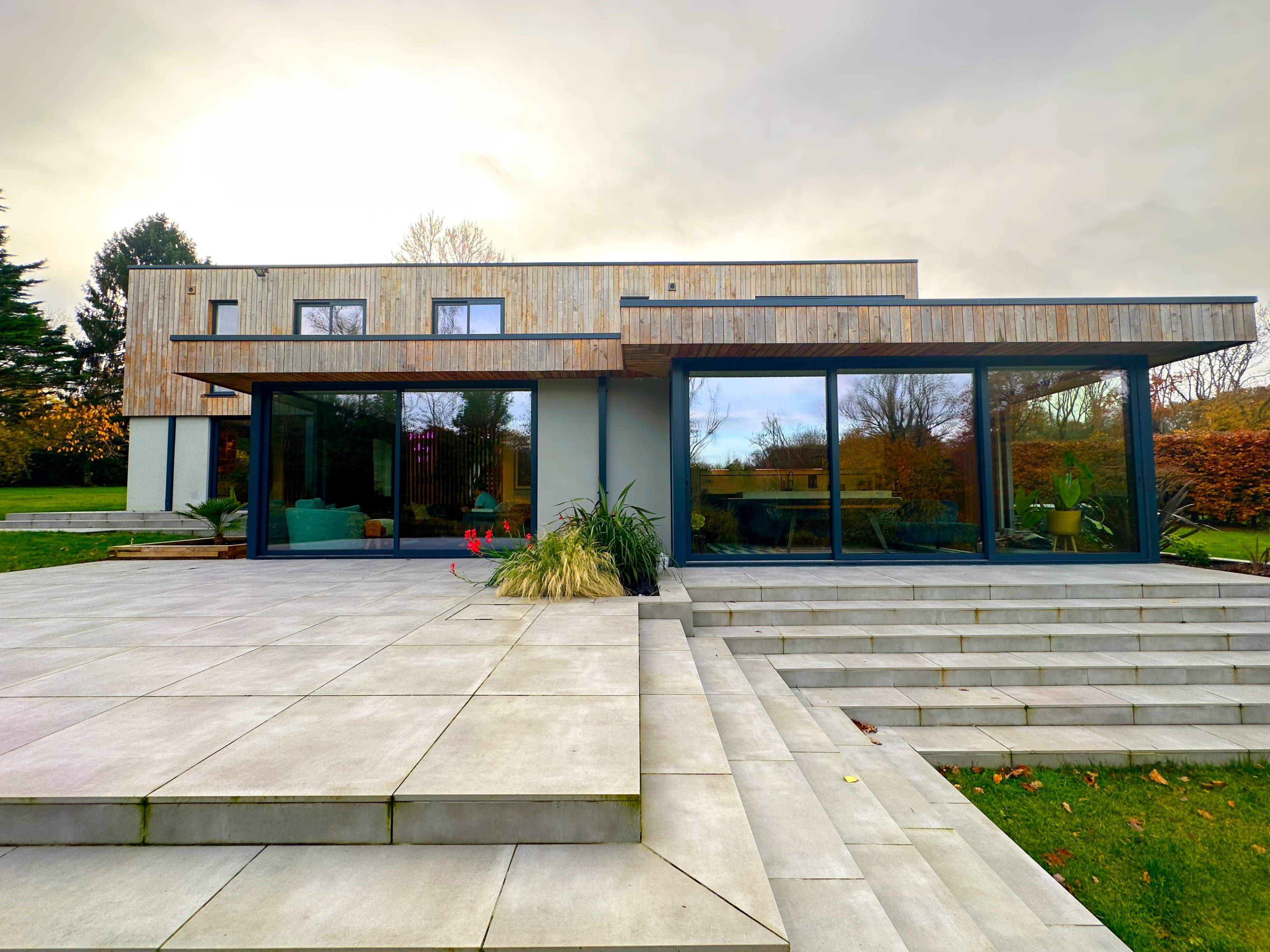 A modern two-story house with a wooden facade, large windows, and a paved patio leading to a well-manicured lawn.