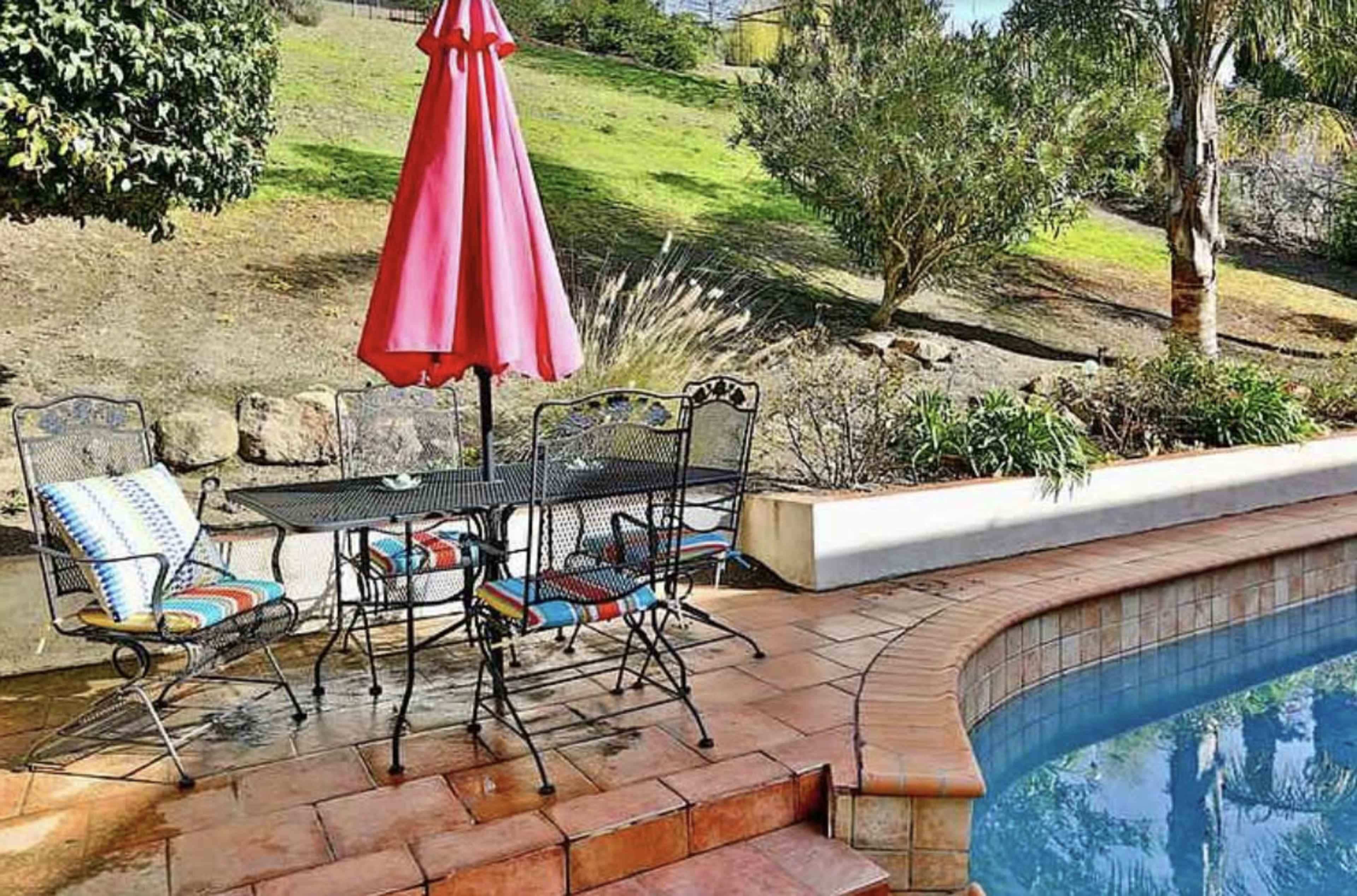 A patio area features a round table with four chairs and a large pink umbrella beside a swimming pool.