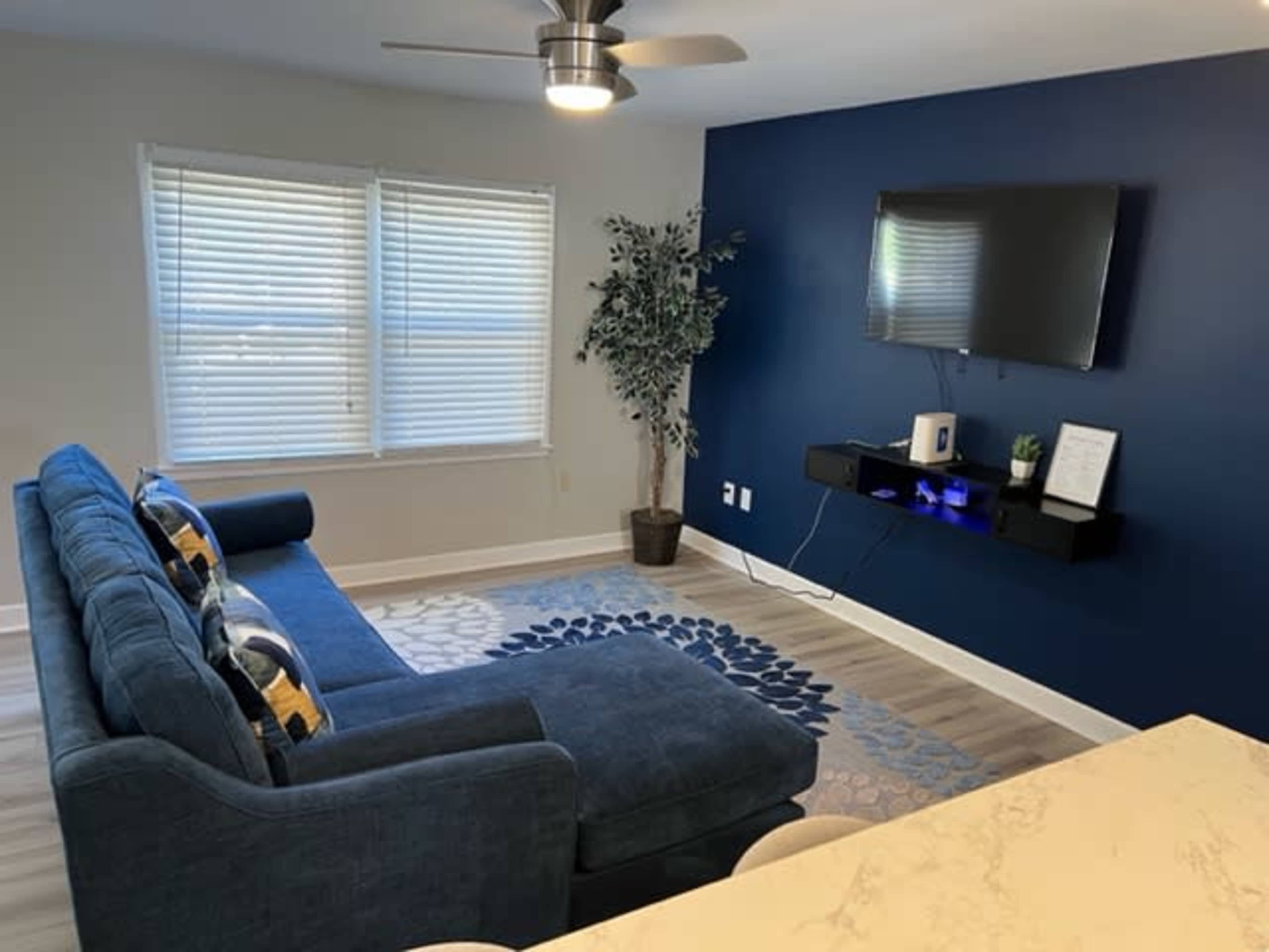 A living room featuring a blue sectional sofa, a wall-mounted TV, and a small shelf with decorative items against a dark blue wall.
