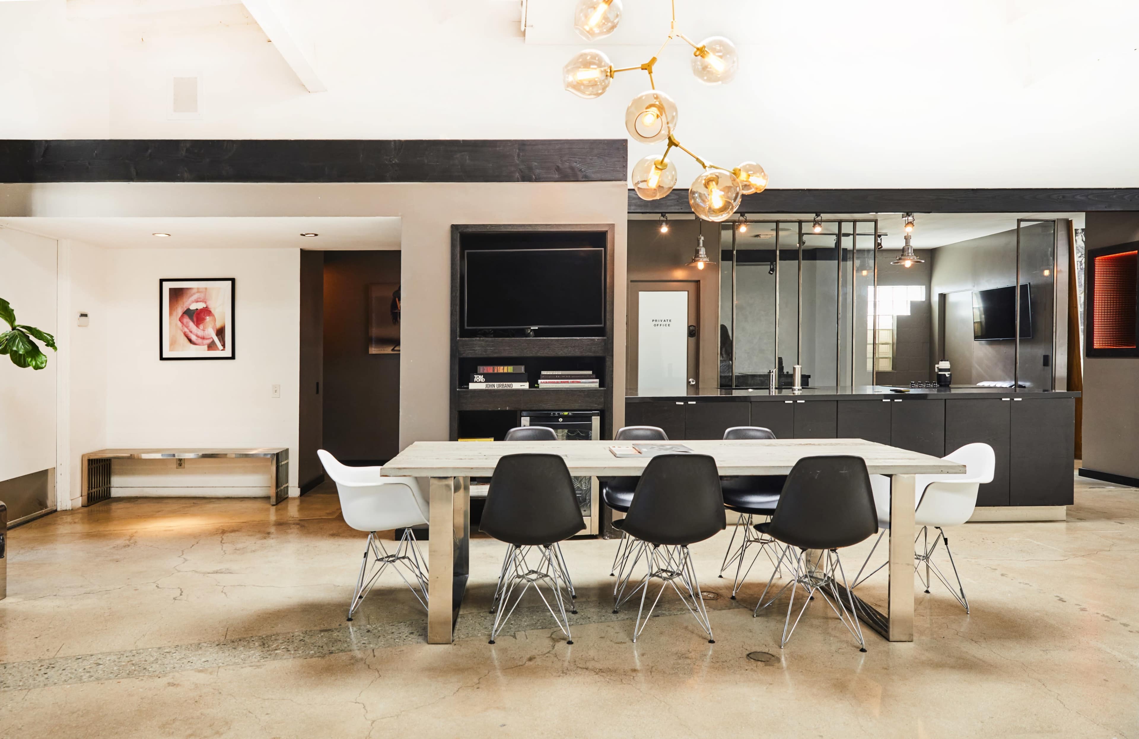 The image shows a modern dining area with a long table surrounded by black and white chairs, illuminated by a pendant light, adjacent to a kitchen area with glass partitions.