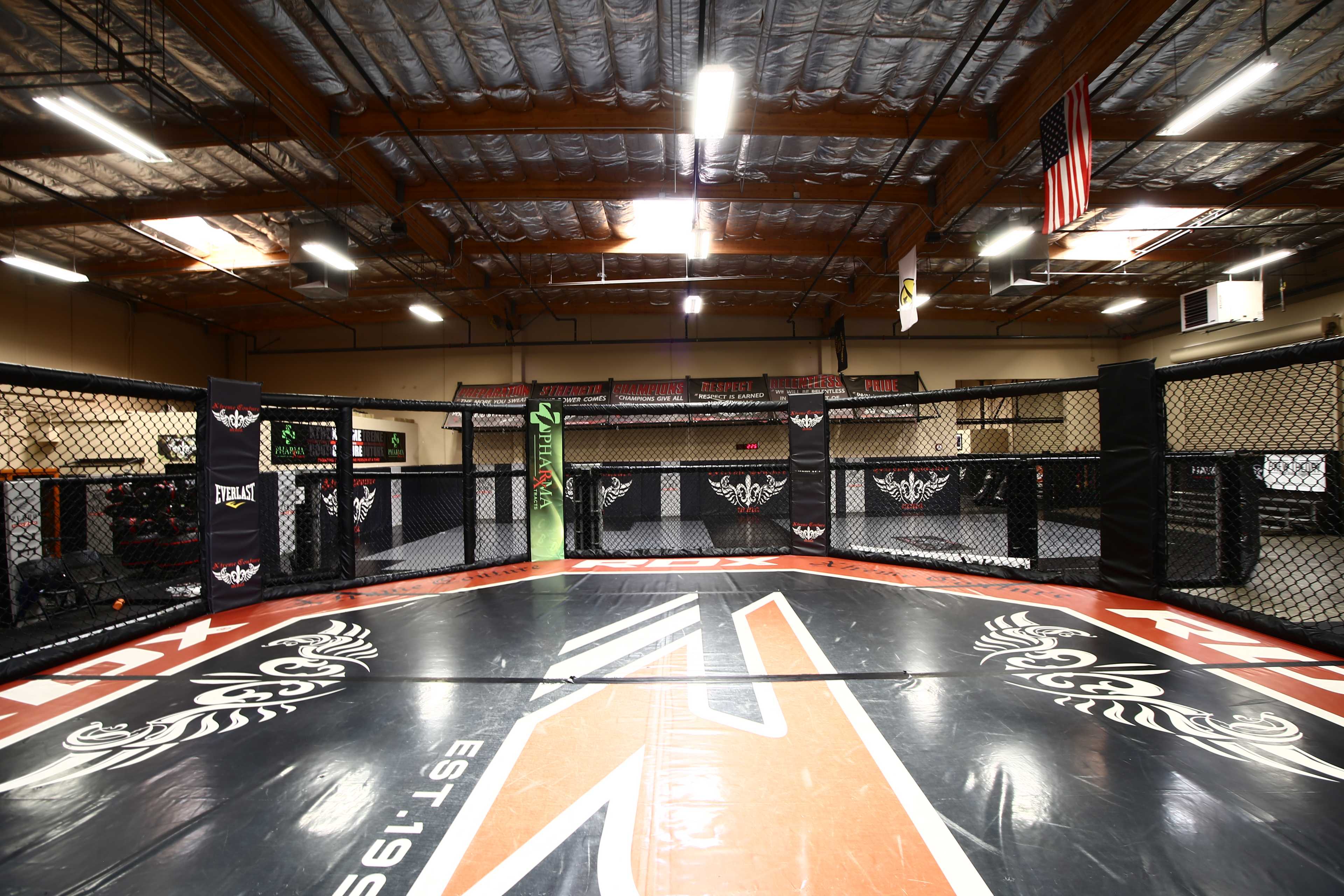 An empty MMA training facility featuring a central octagonal cage surrounded by training equipment and mats.