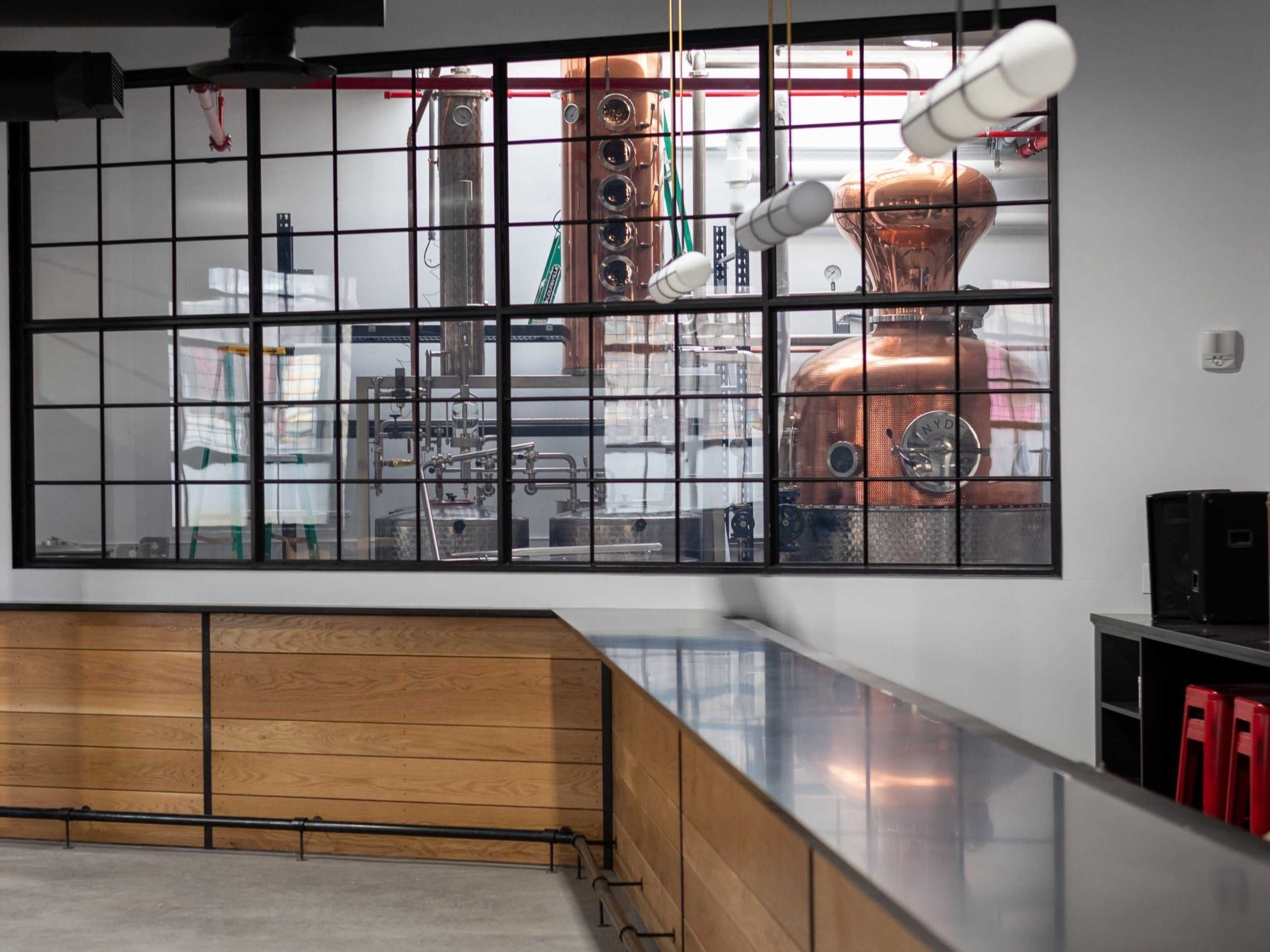 A distillation setup visible through a large glass window in a modern workspace, featuring copper equipment and a bar counter.