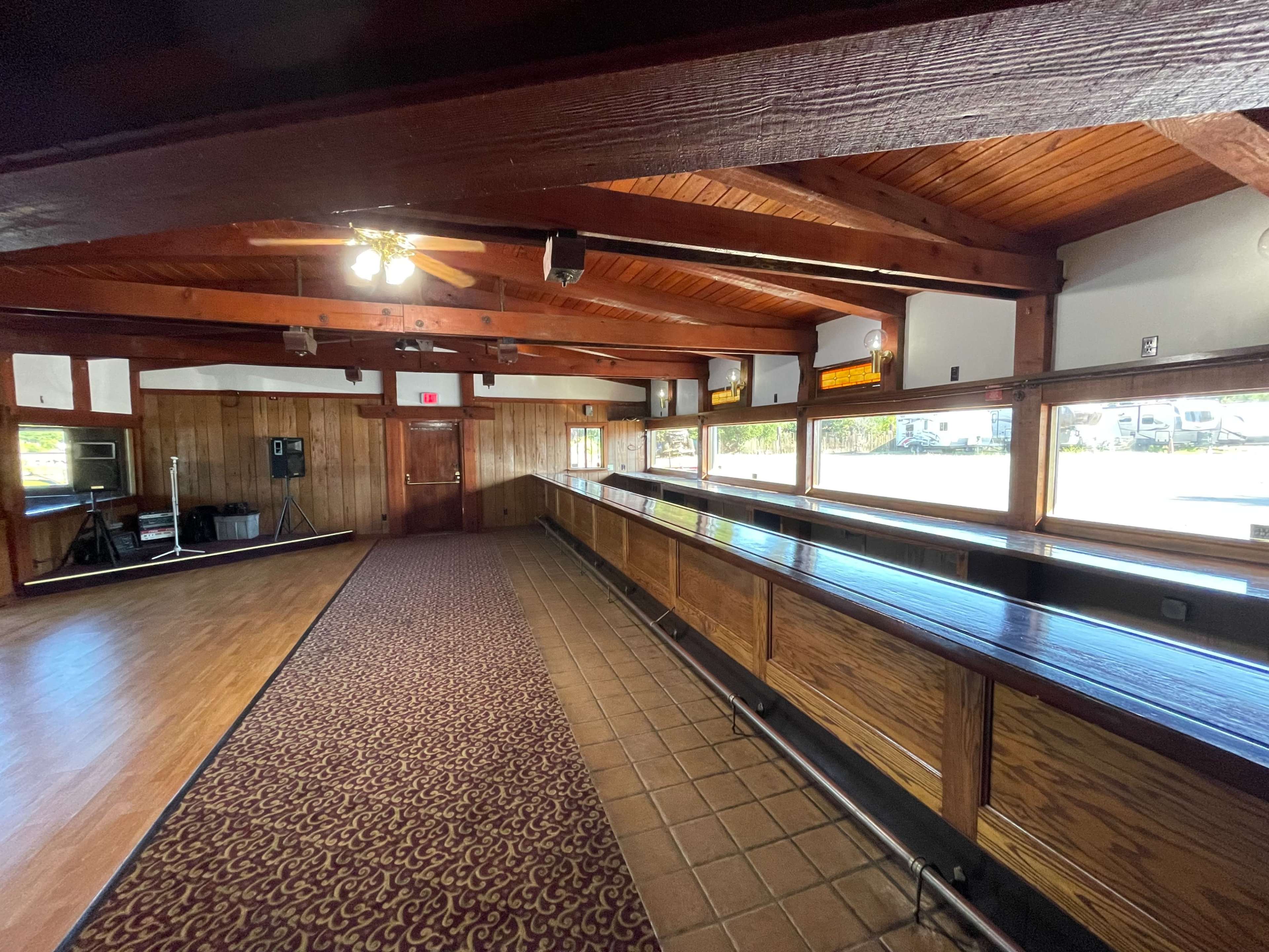 The image shows a spacious interior of a building featuring wooden beams, a long bar area, and a patterned carpet.